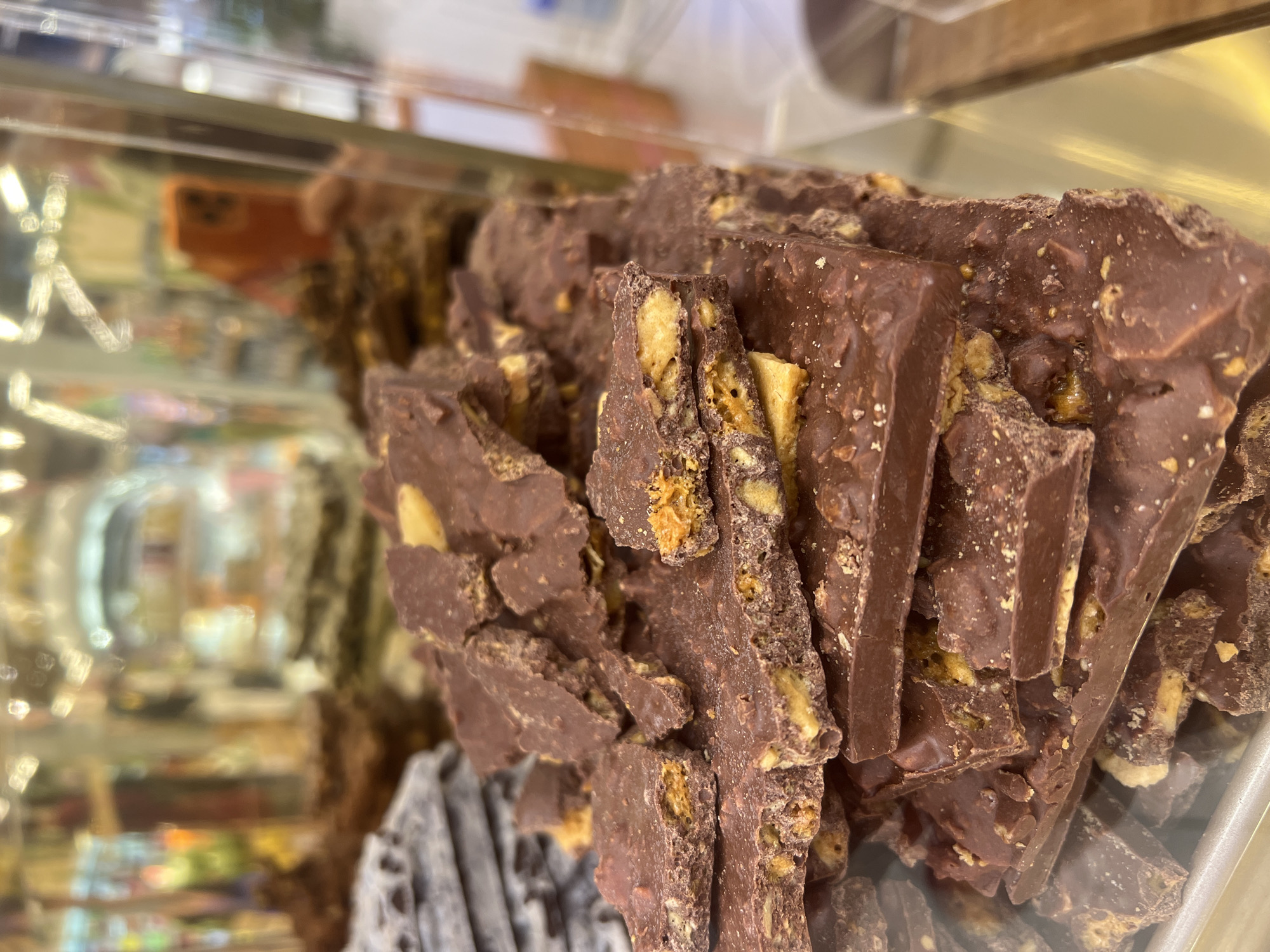 The image displays a close-up, tilted view of numerous pieces of dark chocolate bark, heavily embedded with various nuts and possibly crisps, creating a textured and inviting appearance. The chocolate pieces are irregular in shape and size, suggesting they have been broken from a larger slab, and are piled high within a display.
The chocolate is contained within what appears to be a clear glass or plastic display case, evident from the reflections on its surface. A prominent, blurred reflection of an orange-colored object, possibly a smartphone, is visible in the upper left quadrant, along with other indistinct reflections of ambient light and surroundings.
The background is significantly blurred, indicating a shallow depth of field that keeps the focus sharply on the chocolate. The out-of-focus elements in the background suggest a retail environment, such as a confectionery shop or a market stall, with other items or shelves visible as indistinct shapes and lights. No specific text is visible within the frame. The scene portrays chocolate offered for sale in Denpasar, Indonesia.