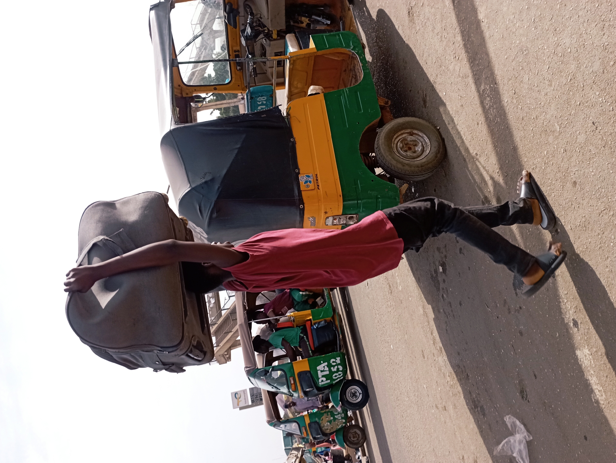 The image captures a vibrant street scene in Jos, Nigeria, featuring a person carrying a large suitcase on their head amidst several auto-rickshaws. The main subject, a young person wearing a red t-shirt, dark trousers, and sandals, stands in the foreground, balancing a large, grey soft-sided suitcase on their head with both arms extended upwards to steady it.
The setting appears to be a busy roadside or a designated area for public transport. Several auto-rickshaws, commonly known as Keke Napep in Nigeria, are lined up behind the main subject. The closest auto-rickshaw is predominantly yellow and green with a black fabric canopy. It displays the word "PETROL" on its side, alongside a blue and white circular logo. A small sign with the number "258" in blue text on a white background is also visible on its front. Further down the line, another auto-rickshaw, also green and yellow, clearly shows the registration number "PTA-1052" on its rear panel. More similar vehicles are visible in the background, suggesting a waiting queue or a transport station.
The ground is a light-colored asphalt road, showing distinct shadows, indicating bright sunlight. A clear, bright sky suggests fair weather. The scene conveys a sense of daily activity and local transportation in an urban or semi-urban environment.