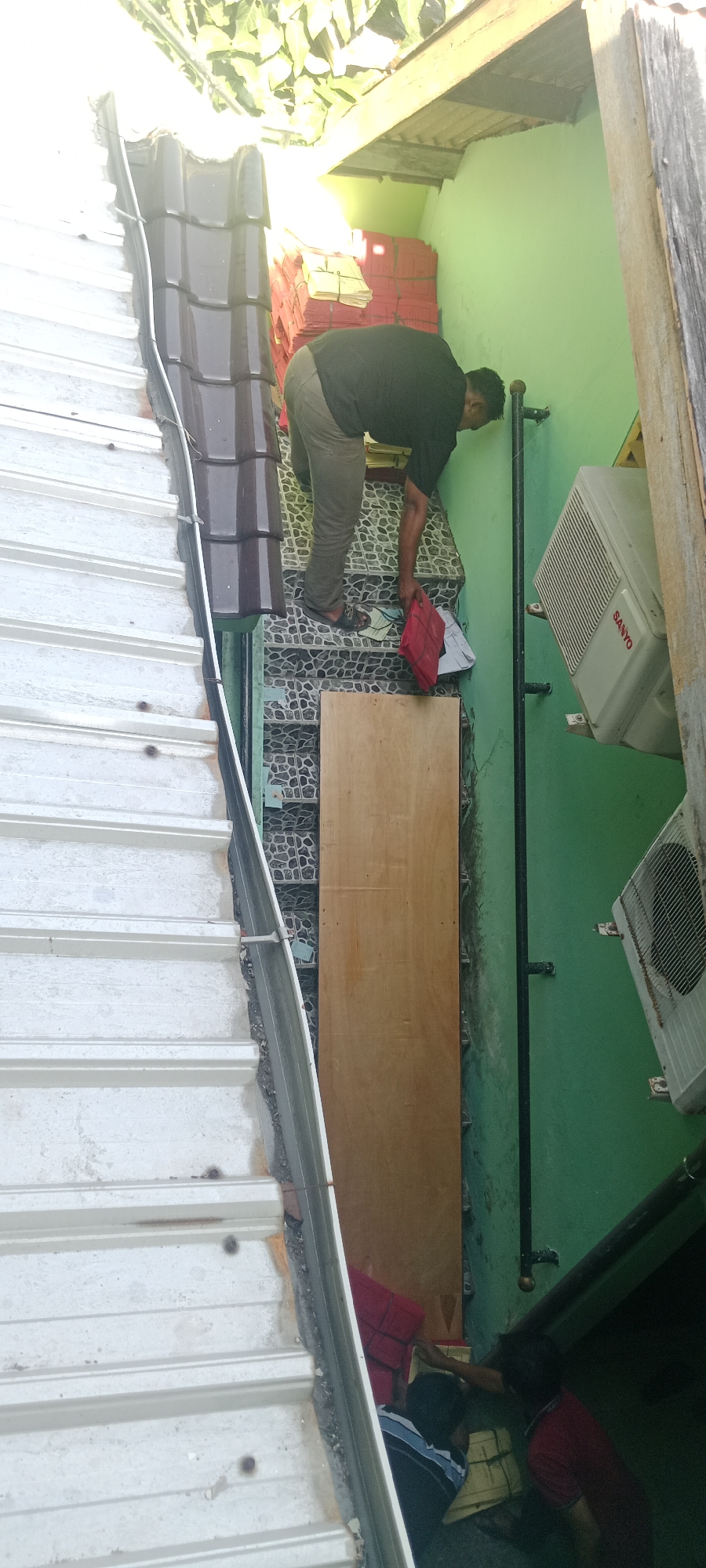 The image depicts an outdoor scene with a person in the upper portion, dressed in dark clothing and gray pants, bent over on a set of stairs. The stairs are partially visible, constructed with a dark tiled pattern. To the left, a corrugated metal structure, possibly a roof or a wall, runs parallel to the stairs. A black pipe is affixed to a light green wall on the right side of the stairs, where an air conditioning unit is mounted. In the lower portion of the image, two other individuals are partially visible, appearing to be working on or interacting with items on the stairs, one wearing a red shirt and the other a blue and white striped shirt. The environment suggests an urban or semi-urban setting, possibly a residential or commercial area in Tuban, Indonesia, given the context. The lighting suggests it is daytime, with some shadows cast.