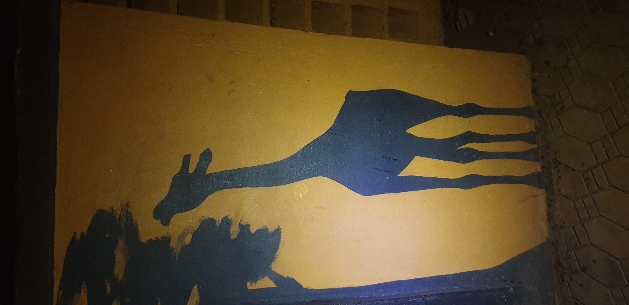 In Narayi, Nigeria, a mural depicts a dark blue silhouette of a giraffe reaching its head towards a dark blue silhouette of a tree branch on a yellow-painted wall. The wall appears to be part of an exterior structure, possibly a building or a retaining wall, with some visible shelving units in the background on the right. The ground in the foreground is paved with hexagonal tiles. The lighting suggests it is either nighttime or an interior space with artificial lighting.