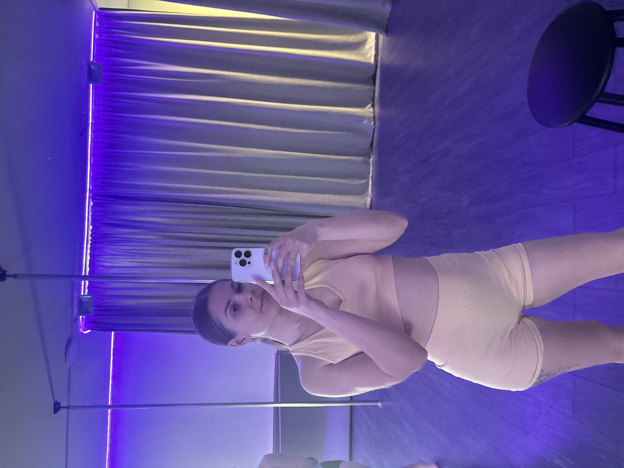 A young woman is captured in a full-length selfie, taken indoors in what appears to be a modern dance or fitness studio in Batumi, Georgia. The image is presented rotated 90 degrees counter-clockwise.
The woman, with dark hair pulled back, is wearing a ribbed, light yellow or cream-colored two-piece athletic outfit, comprising a sleeveless crop top and high-waisted biker shorts. She holds a light-colored smartphone in her right hand, taking the photograph with a neutral expression, looking directly towards the phone's lens.
The background features light-colored curtains along one wall, illuminated by a warm, yellowish light from above. Adjacent to these curtains, a bright purple LED strip light runs vertically along the wall, casting a dominant purple glow throughout the room. A small black electrical box is mounted on the wall near the top of this LED strip. Another purple LED strip is visible on the floor, and the floor itself, possibly light-colored wood or tile, reflects the purple illumination.
To the left of the woman, a gleaming metal pole, characteristic of a pole dancing or fitness studio, extends from the floor to the ceiling. A black stool with a round seat is partially visible on the right side of the frame. The artificial and colored lighting creates a distinct, contemporary ambiance, suggesting the photo was taken either at night or in a purposefully lit space. No discernible text is visible in the image.