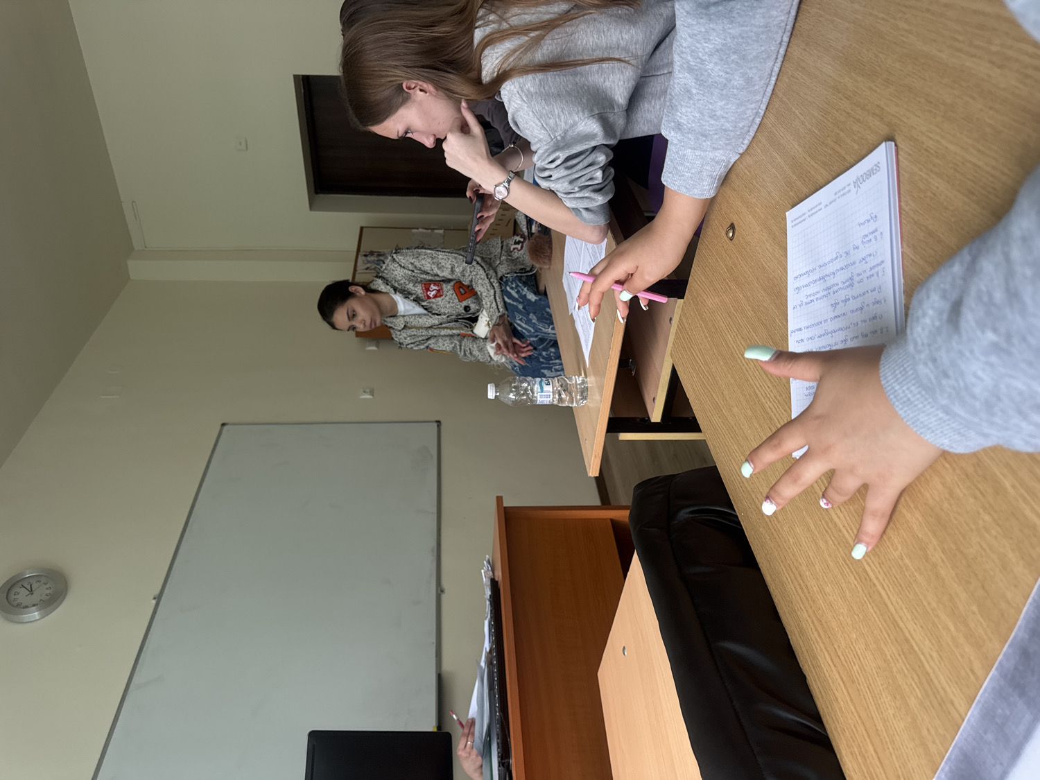 An interior shot depicts a classroom or study area with several young women engaged in an activity. The room features light-colored walls, wooden desks, and a white magnetic whiteboard. Natural light illuminates the space.
In the foreground, the hands and forearms of a person wearing a grey sweatshirt are visible, resting on a light-colored wooden desk. This person has light green nail polish on several fingers, with some nails featuring white accents. To their left, a white notebook is open on the desk, displaying handwritten text in Cyrillic script and the word "SEMBROOK" on its cover. The text includes the heading "Задачи" (Tasks) and numbered points, suggesting notes or assignments. A pink pen is held by another hand, also wearing a grey sweatshirt, belonging to a second person seated further back at the same desk. This individual has long brown hair and is looking down intently at something held in their hands, possibly a mobile device, though it's obscured. They are wearing a watch on their left wrist.
Further into the background, to the left, a third woman with dark hair tied back is seated at another desk. She wears a patterned jacket and blue jeans and is holding a clear plastic water bottle. Her gaze is directed downwards. On the left wall, a round silver clock shows the time as approximately 12:55. A dark wooden door frame or cabinet is visible in the background, to the right of the whiteboard. The overall atmosphere suggests a focused, academic, or collaborative setting during the daytime.