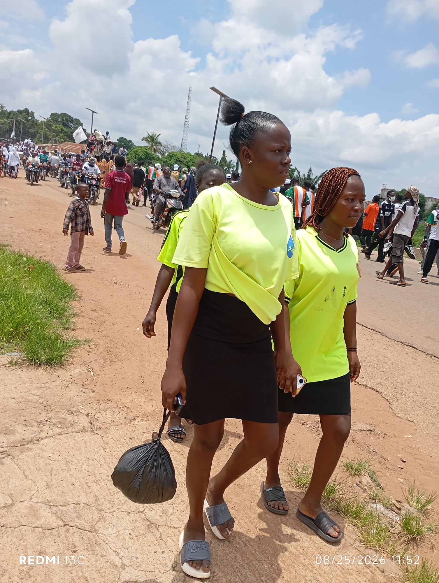 In Shendam, Nigeria, two young women in neon green t-shirts and black skirts walk side-by-side on a dusty, unpaved road. The woman on the left, with her hair in a bun, carries a black plastic bag in her right hand. The woman on the right has braided hair. In the background, a crowd of people, many on motorcycles, are visible. The sky is partly cloudy. The scene appears to be a procession or parade.