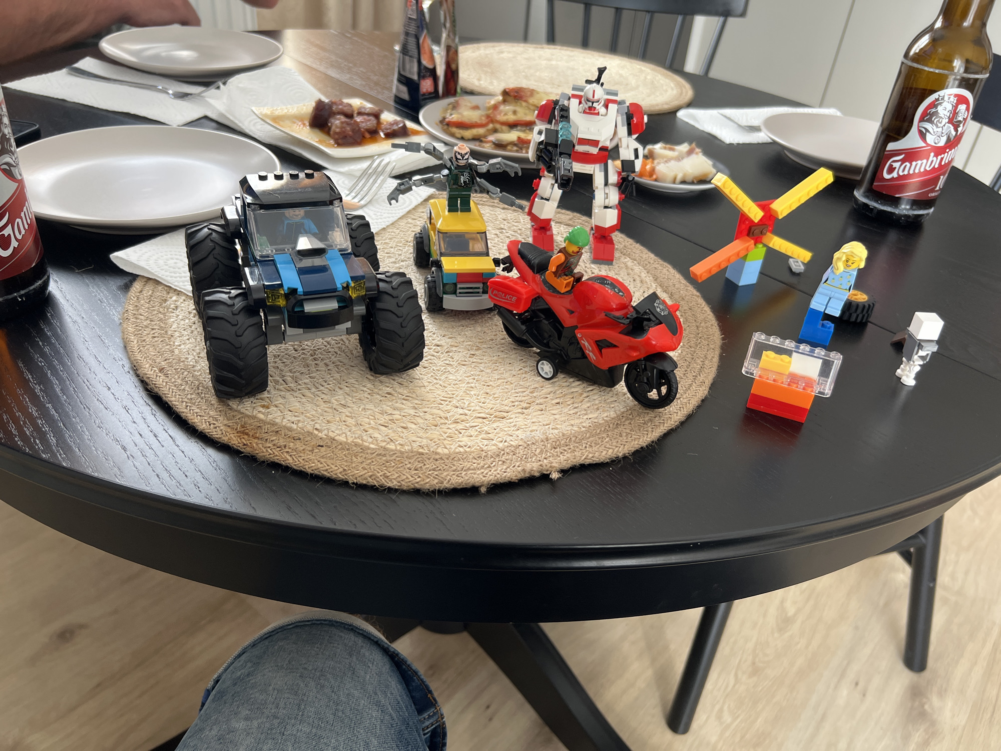 This media file is a photograph taken indoors, likely at a dining table, capturing a collection of LEGO toys alongside food and drinks.
The subjects include several LEGO creations: a large blue and black monster truck with a minifigure driver, a smaller yellow vehicle with a green minifigure with four arms standing on top, a red police motorcycle with a minifigure rider, and a large red and white robot minifigure holding a blue weapon. There are also smaller LEGO elements scattered around, including a colorful windmill-like structure, a minifigure of a blonde woman with blue attire and boots, and some loose blocks and a small white LEGO piece.
On the table, partially visible are two white plates, one with some food that appears to be meat in sauce, and another with what looks like slices of bread or a similar baked item topped with cheese or a white sauce. A bottle of Gambrinus beer is prominently displayed, with its label clearly visible. The setting is a dark, round dining table with a woven placemat under some of the LEGO figures. In the background, parts of chairs and possibly other people's hands are visible, suggesting a mealtime or gathering. The floor appears to be light-colored wood laminate.
The overall mood seems casual and perhaps playful, with the LEGO toys arranged as if in a scene. There are no obvious cues about the time of day or weather. The location context is Budapest, Hungary.
Visible text includes "Gambrinus" on the beer bottle.