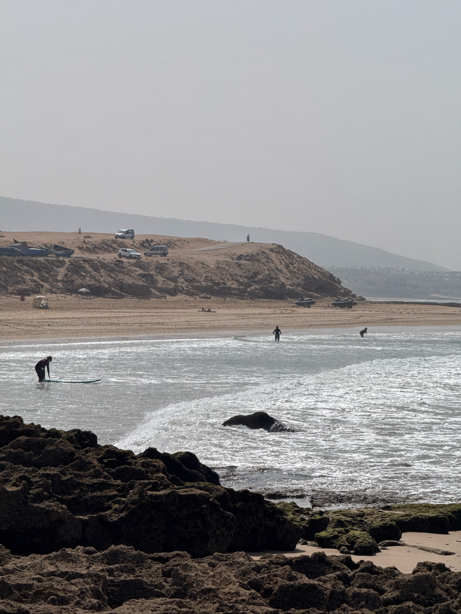 This is an image of a beach in Aourir, Morocco. In the foreground, rough, dark brown rocks and sand are visible. The middle ground shows a sandy beach with a few scattered objects including a small tent, an umbrella, and what appear to be several boats pulled up on the sand. Further up the embankment, there are several parked vehicles including two white vans and one white SUV. There are also several other smaller vehicles visible on the sandy area. The background consists of a sandy hill with a road winding along its crest, leading to a hazy, mountainous landscape. In the water, there are a few individuals. On the left, a person is standing in the water with a surfboard. Further out in the water, another person is visible, also near a surfboard. On the far right, a person is bent over in the shallow water. The water itself is choppy with sunlight glinting off the surface. The sky is overcast and hazy.