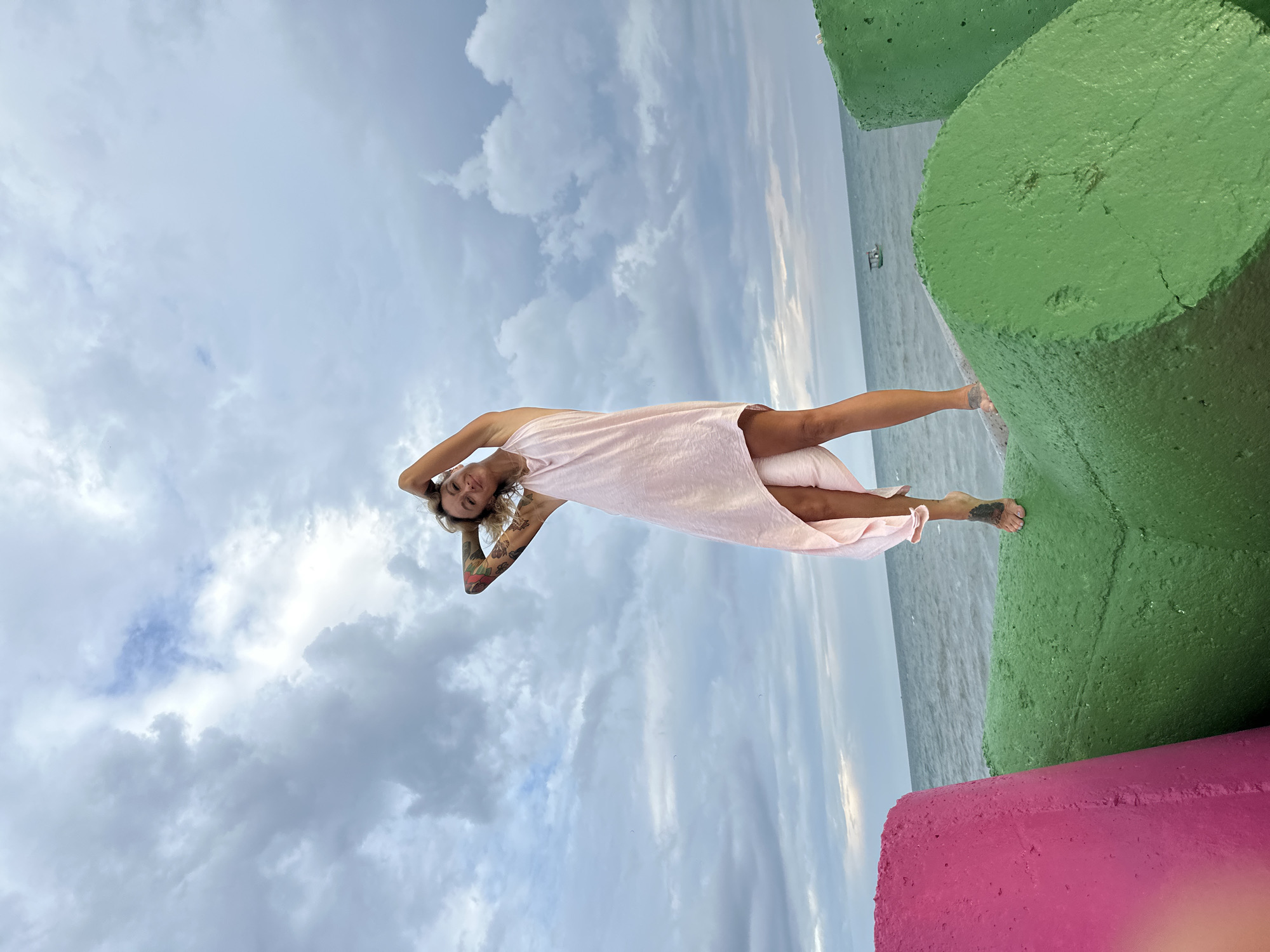 The image shows a woman posing on a colorful concrete breakwater by the sea, under a cloudy sky. The woman, with blonde hair, is wearing a light pink, flowing dress or cover-up. She is standing with her arms raised behind her head, looking towards the camera with a relaxed smile. Tattoos are visible on her arms and left foot.
To her right, the calm, grey-blue sea stretches towards the horizon, with a small boat visible in the distance. In the foreground and extending to the right, large, textured concrete breakwater blocks are painted in vibrant green and pink. The sky above is largely covered with white and grey clouds, suggesting an overcast day. The scene captures a moment of leisure in a coastal setting. There is no visible text.