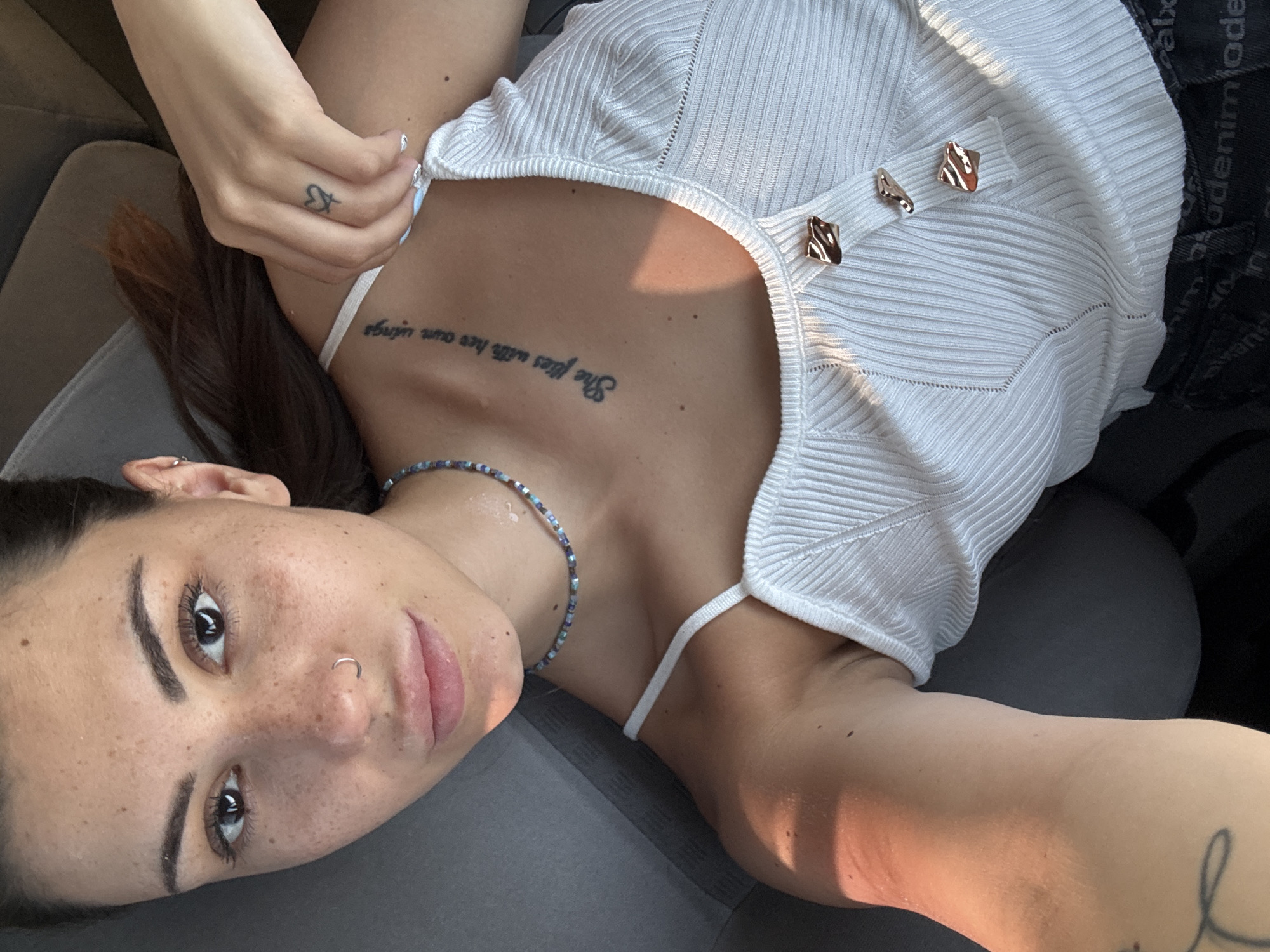 The media file shows a young woman lying down, with her head tilted towards the camera. She has long brown hair, dark eyes, and visible freckles on her face and arms. She is wearing a white ribbed tank top with decorative gold buttons and a beaded necklace. A tattoo of text is visible on her chest, reading "She flies with her own wings." Another tattoo, a small heart with an arrow through it, is on her left hand. She also has a nose ring. The lighting suggests it is daytime, with warm sunlight casting shadows. The setting appears to be indoors, possibly in a car, as indicated by the upholstered seat. There are no overt signs of weather conditions. The woman's expression is neutral. Visible text on dark fabric behind the woman reads "denim mode".