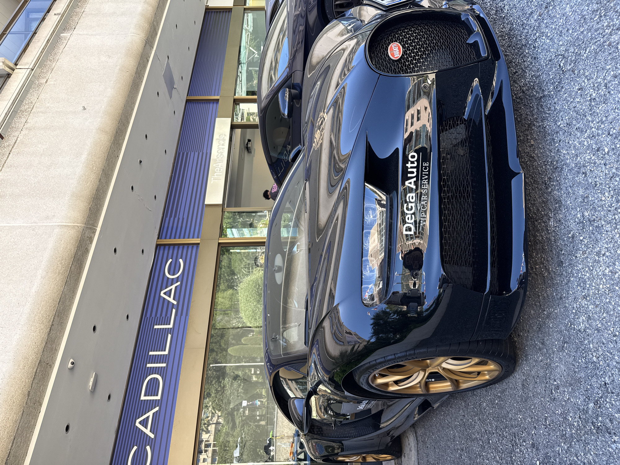 A black Bugatti Chiron Super Sport 300+ is parked on a city street in front of a building with "CADILLAC" signage. The car has gold-colored wheels and a prominent Bugatti emblem on the front grille. A sticker on the front bumper reads "DeGa Auto VIP Car Service." To the right of the Bugatti, a dark blue Mercedes-Benz is partially visible. The building facade features large windows and architectural elements typical of a commercial area. The environment appears to be sunny, and the street is paved with asphalt. There are no people clearly interacting with the cars or in the immediate foreground. The location is identified as Monaco, Monaco.