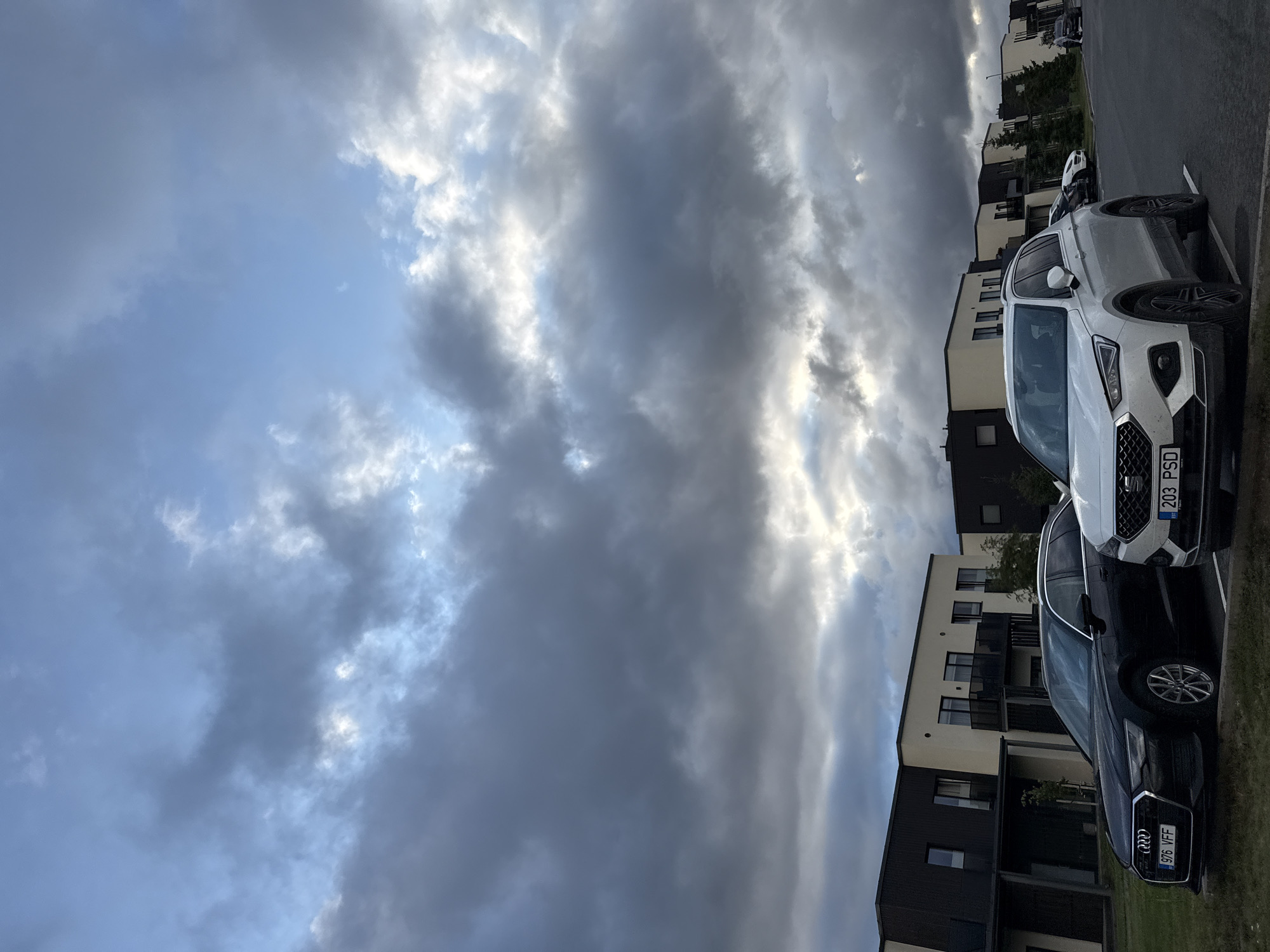 The image captures a street scene in Peetri alevik, Estonia, under a dramatic sky filled with dark, heavy clouds interspersed with patches of bright sunlight. In the foreground, two cars are parked. The car on the left is a black Audi wagon with license plate "976 VFF." The car on the right is a white SUV, possibly a SEAT, with license plate "203 PSD." Behind the cars, a row of modern, multi-story residential buildings with dark and light facades is visible. The architecture is contemporary, featuring balconies and multiple windows. Trees, some appearing to be coniferous, are planted along the sides of the buildings and in front of them. The ground is a mixture of asphalt and grassy areas. The overall atmosphere suggests an overcast day with intermittent sunlight.