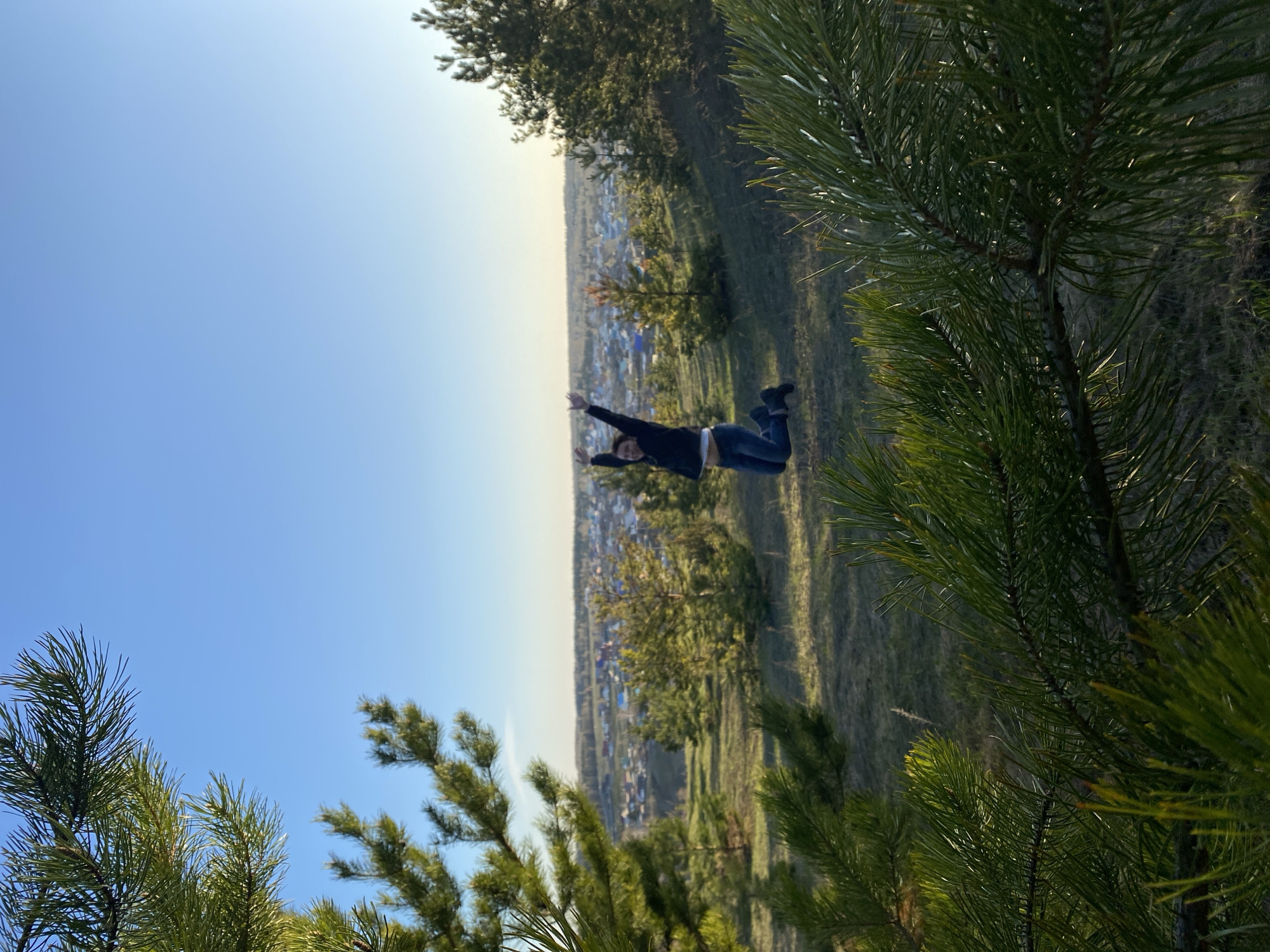 The image depicts a person lying on a grassy hillside, with their arms outstretched. In the background, a town, likely Ольховка, Russia, is visible. The foreground features pine trees framing the shot. The sky is clear and blue. The person is wearing a dark jacket and jeans.