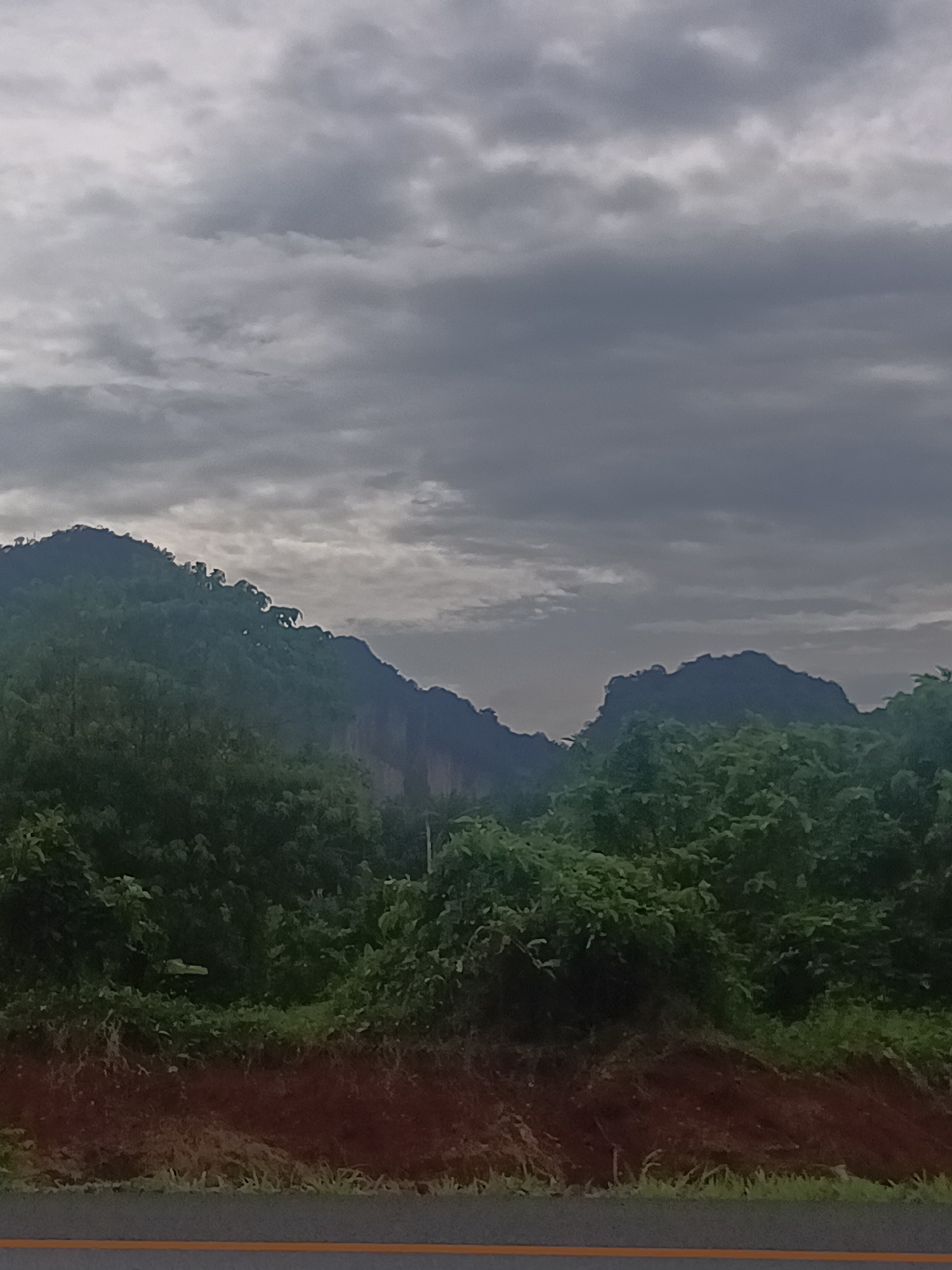 The image depicts a mountainous landscape under a cloudy sky, likely in Plai Phraya, Thailand, based on the context. The foreground shows a road with a yellow line marker. Above the road, there's a brown dirt bank leading to dense green vegetation. A mountain range is visible in the background, with trees covering the slopes. The sky is overcast with heavy, gray clouds. There are no people visible in the image.