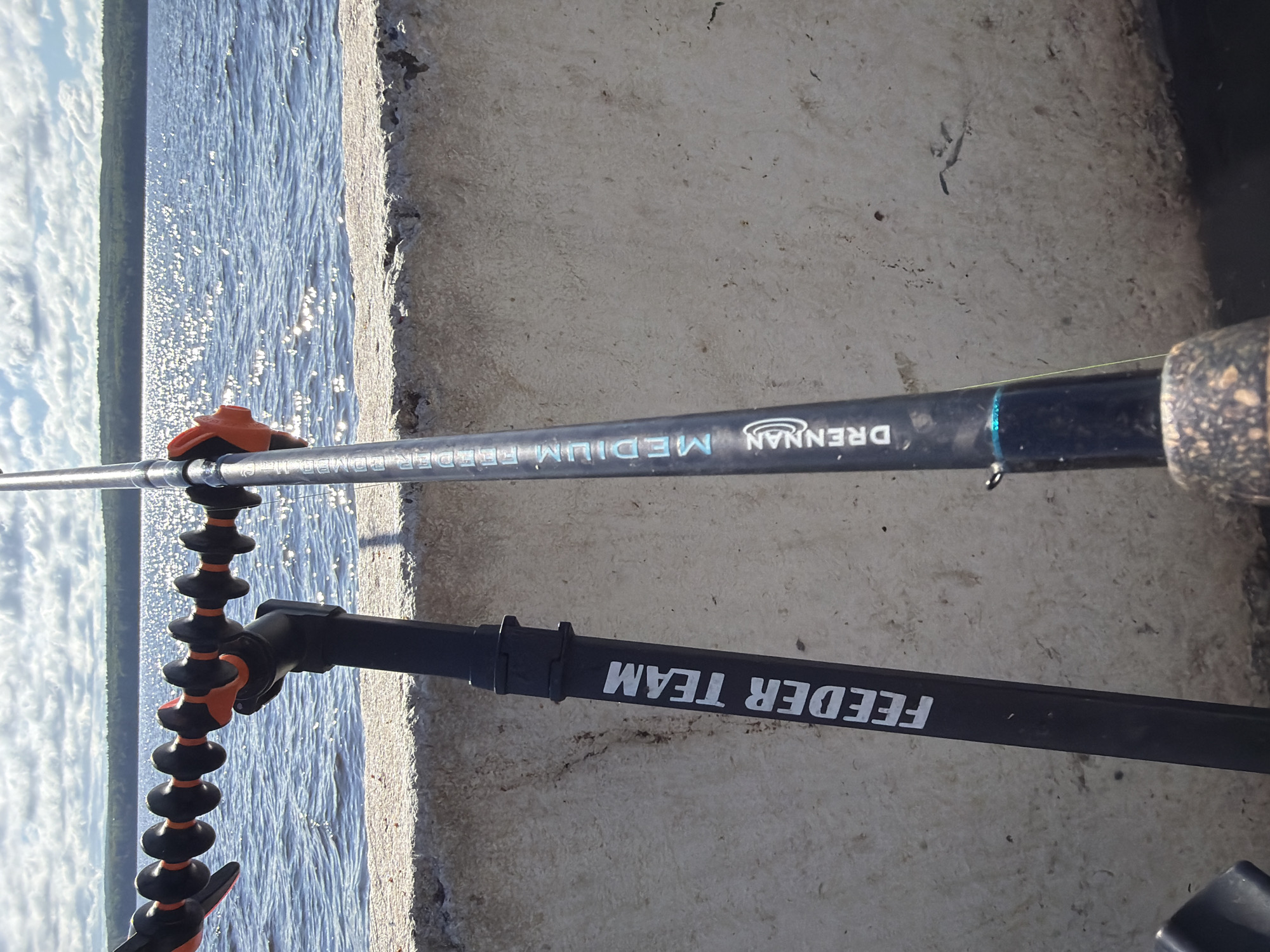 The image displays two fishing rods leaning against a concrete barrier overlooking a body of water. The rod on the left has "FEEDER TEAM" written on its black handle. The rod on the right has "DRENNAN MEDIUM FEEDER" written on its dark blue handle. A flexible rod holder with black and orange segments is attached to the fishing rod on the right, securing it. The water reflects the sunlight, and a treeline can be seen in the distance. The sky is partly cloudy. The setting appears to be an outdoor location, possibly a pier or waterfront. The city of Chaikovsky, Russia, is not depicted in this image.