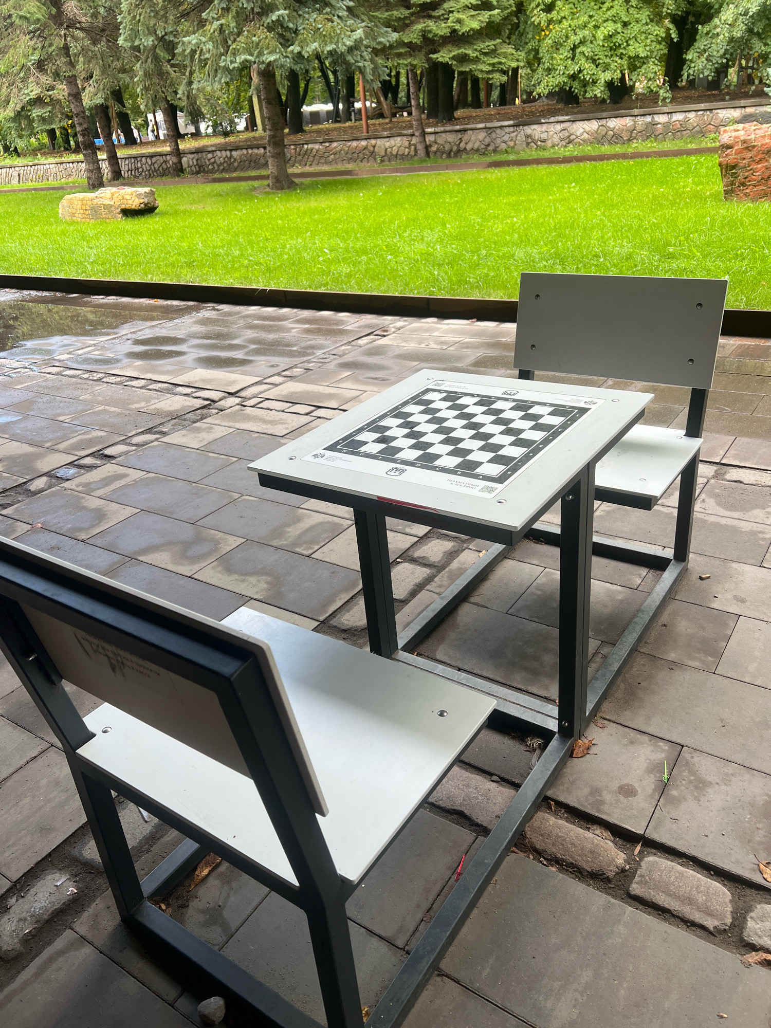 The image depicts an outdoor chess table with two white chairs on a paved area in front of a grassy park. The chess board is set up on the table, and the chairs appear to be made of metal and white material. The ground is tiled with gray stones, some of which are wet, indicating recent rain. In the background, there are trees, a stone wall, and a lush green lawn. The overall setting suggests a public park or recreational area. The location is identified as Kaliningrad, Russia.