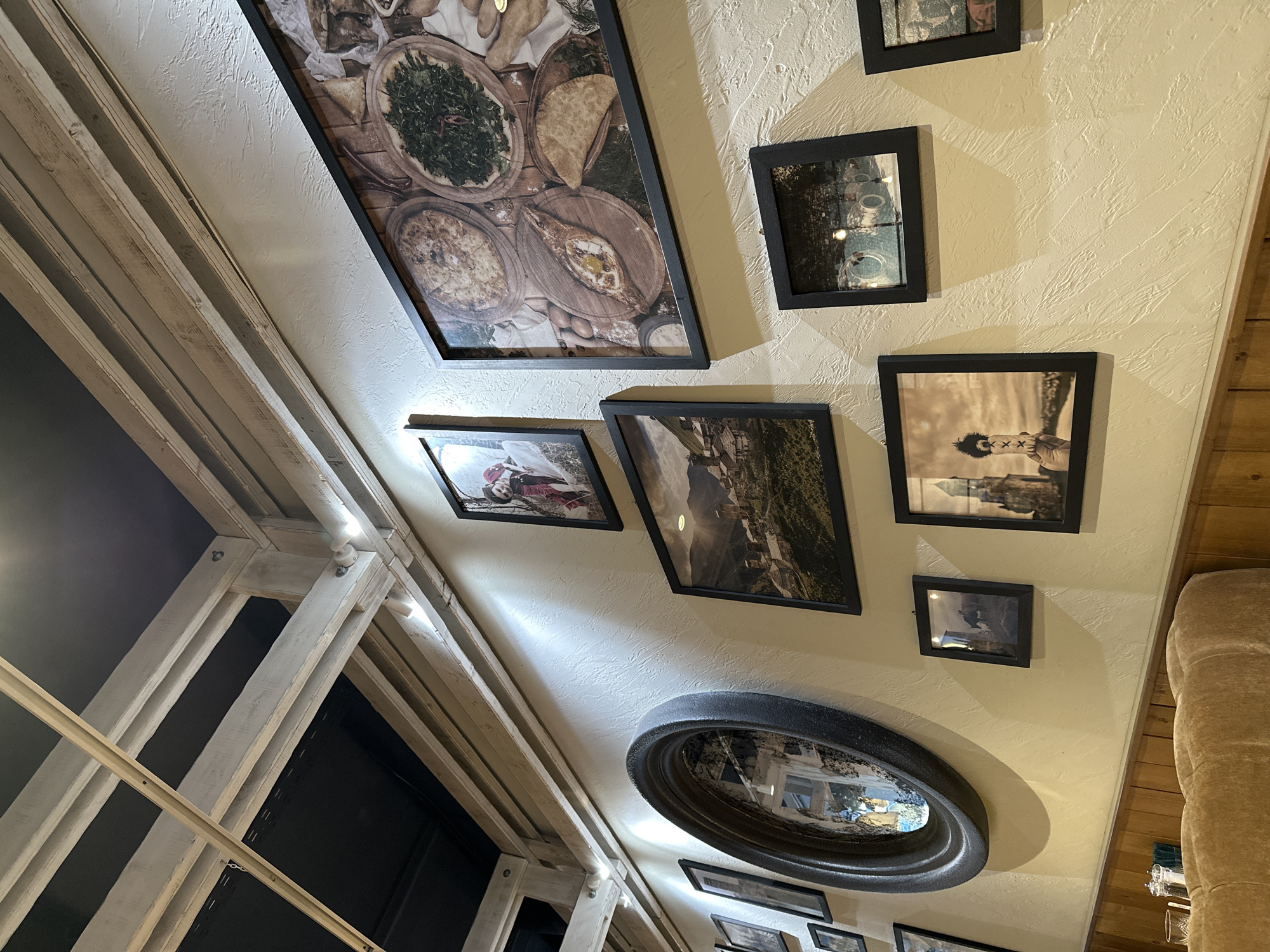 The image shows a wall decorated with several framed pictures and a mirror. The pictures vary in size and depict different scenes, including food, landscapes, and portraits. The mirror is oval-shaped with a dark frame. A window with a white frame is visible on the left side of the image. The wall is painted in a light color with a textured finish. A portion of a beige sofa is seen on the right. There is no indication in the image itself that it is located in Izhevsk, Russia.