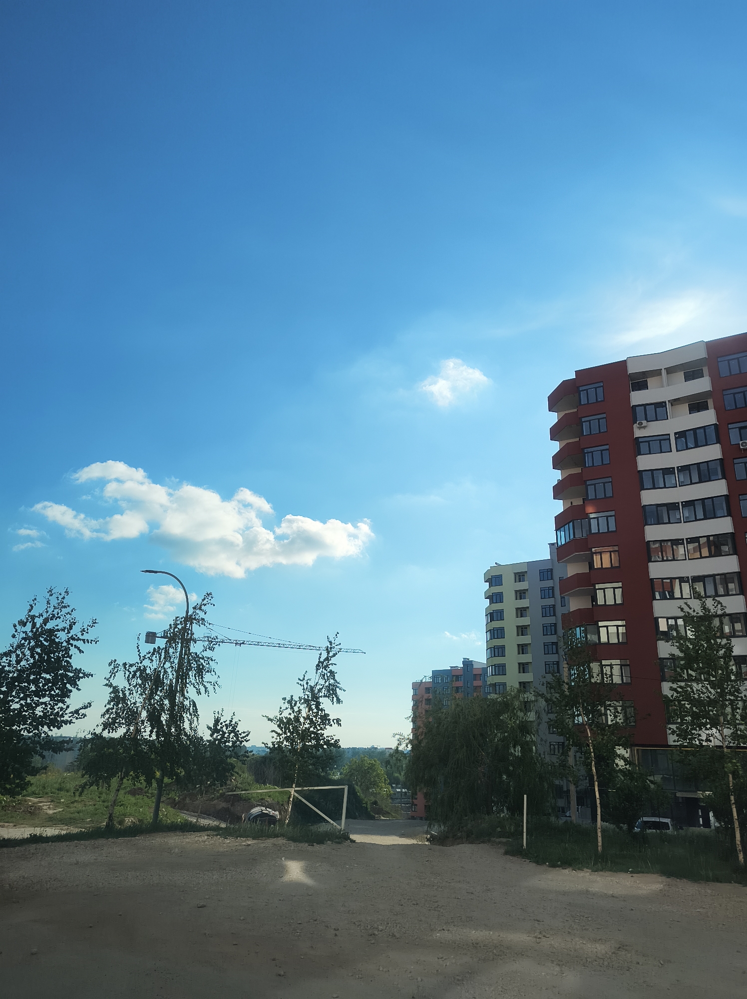 The image shows a residential area in Ternopil, Ukraine, under a blue sky with scattered clouds. In the foreground is an unpaved road leading towards a construction site. To the left of the road are sparse trees and overgrown vegetation, with a partially collapsed wooden fence. In the background, a construction crane is visible. To the right, there are newly constructed high-rise apartment buildings of varying colors (red, white, blue, green, grey) and heights, partially obscured by trees. The image appears to have been taken on a sunny day. There are no people visible.