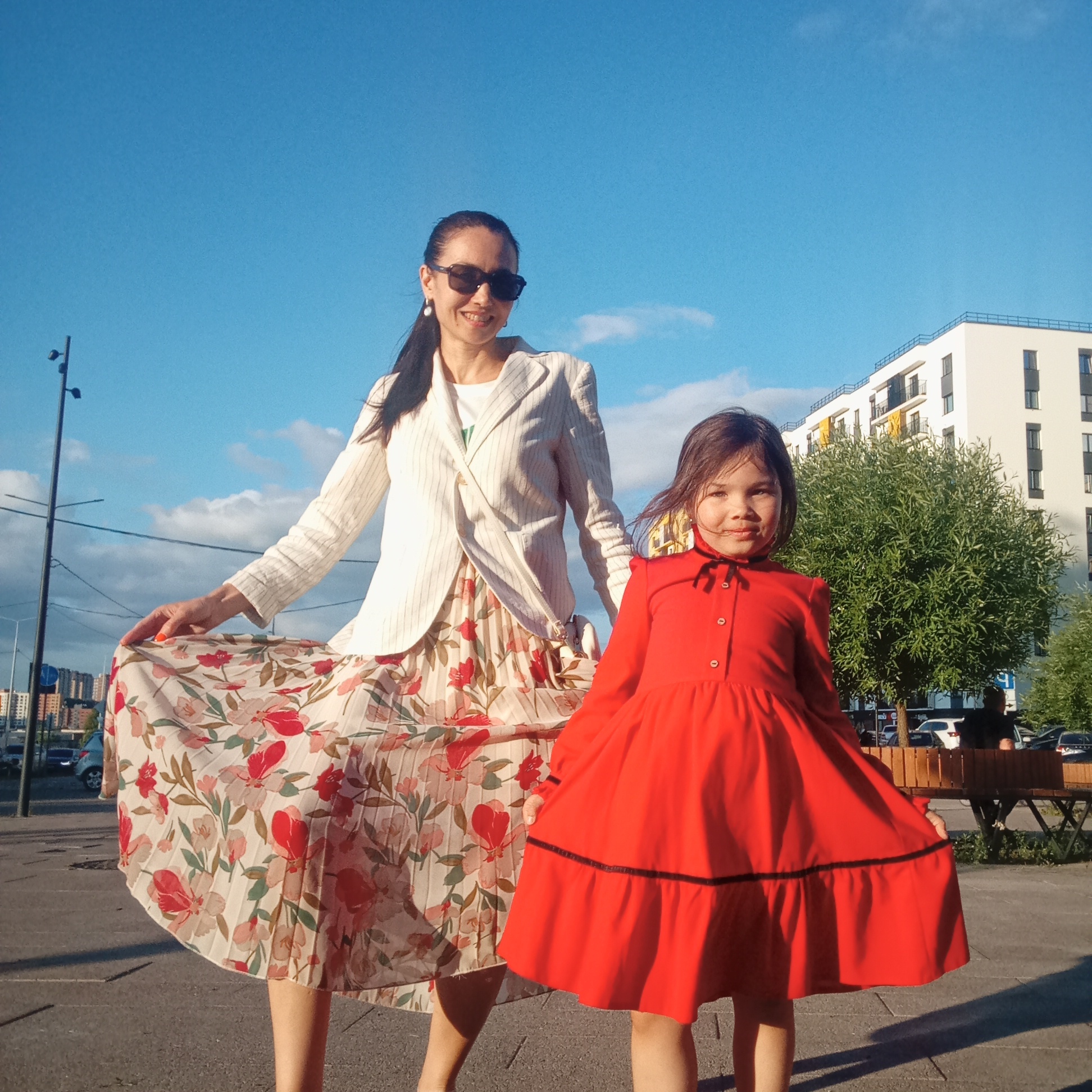 An adult woman and a young girl are captured posing outdoors on a sunny day in Pervomaysky, Russia. The woman, positioned on the left, wears a white striped blazer over a white t-shirt, paired with a long, pleated floral skirt featuring red, pink, and green patterns on a cream background. She has dark hair pulled back, sunglasses, and pearl drop earrings, and is seen smiling while lifting her skirt slightly. Her fingernails have orange polish. To the right, a young girl wears a bright red long-sleeved dress with a black ribbon tied at the collar and a black stripe above the ruffled hem. She also holds her dress out gently and displays a subtle smile.
The scene is set in an urban or suburban environment under a clear blue sky with scattered white clouds. The background includes modern white buildings with dark windows and balconies, green trees, street lights, and parked cars. A wooden bench is visible behind the girl. The lighting suggests it is daytime, possibly late afternoon or early evening, creating some visible shadows. Both subjects appear happy and relaxed. No discernible text is visible on any objects or signs within the image.