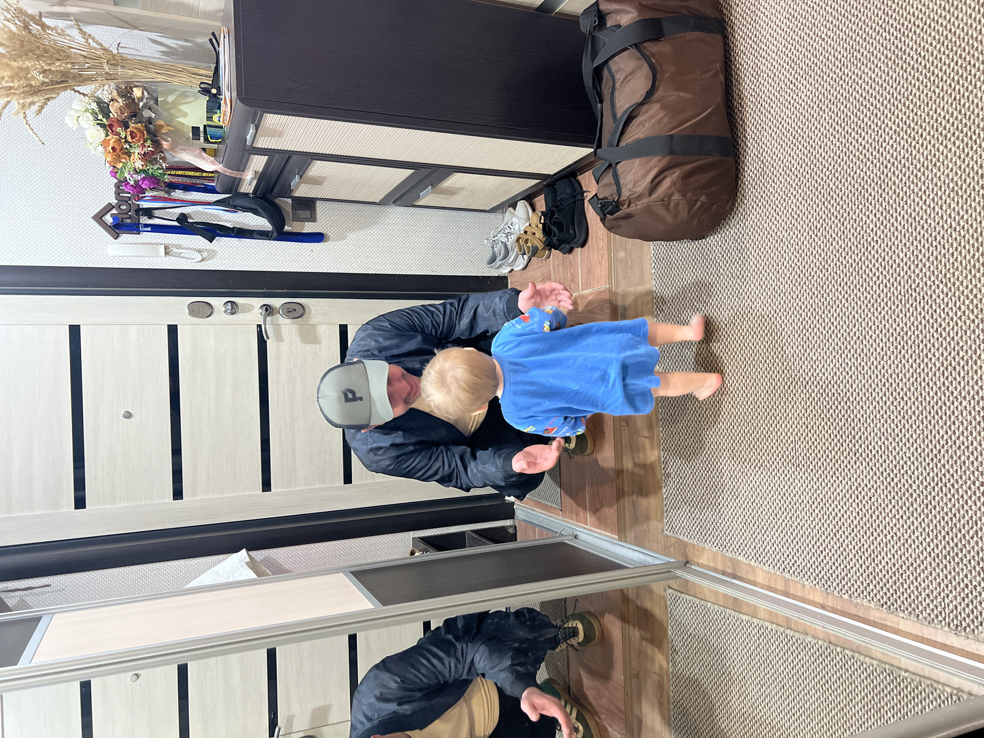 A man wearing a black jacket and grey cap is squatting near a toddler in a blue shirt in what appears to be an entryway. The entryway includes a brown cabinet with drawers, a brown duffel bag, several pairs of shoes, and a mirror. There is a vase of flowers and wheat stalks on top of the cabinet, along with what appears to be a collection of sports medals. The entryway has a beige rug and light-colored wooden floors. It is likely the city Noviy is New Urengoy, Russia.