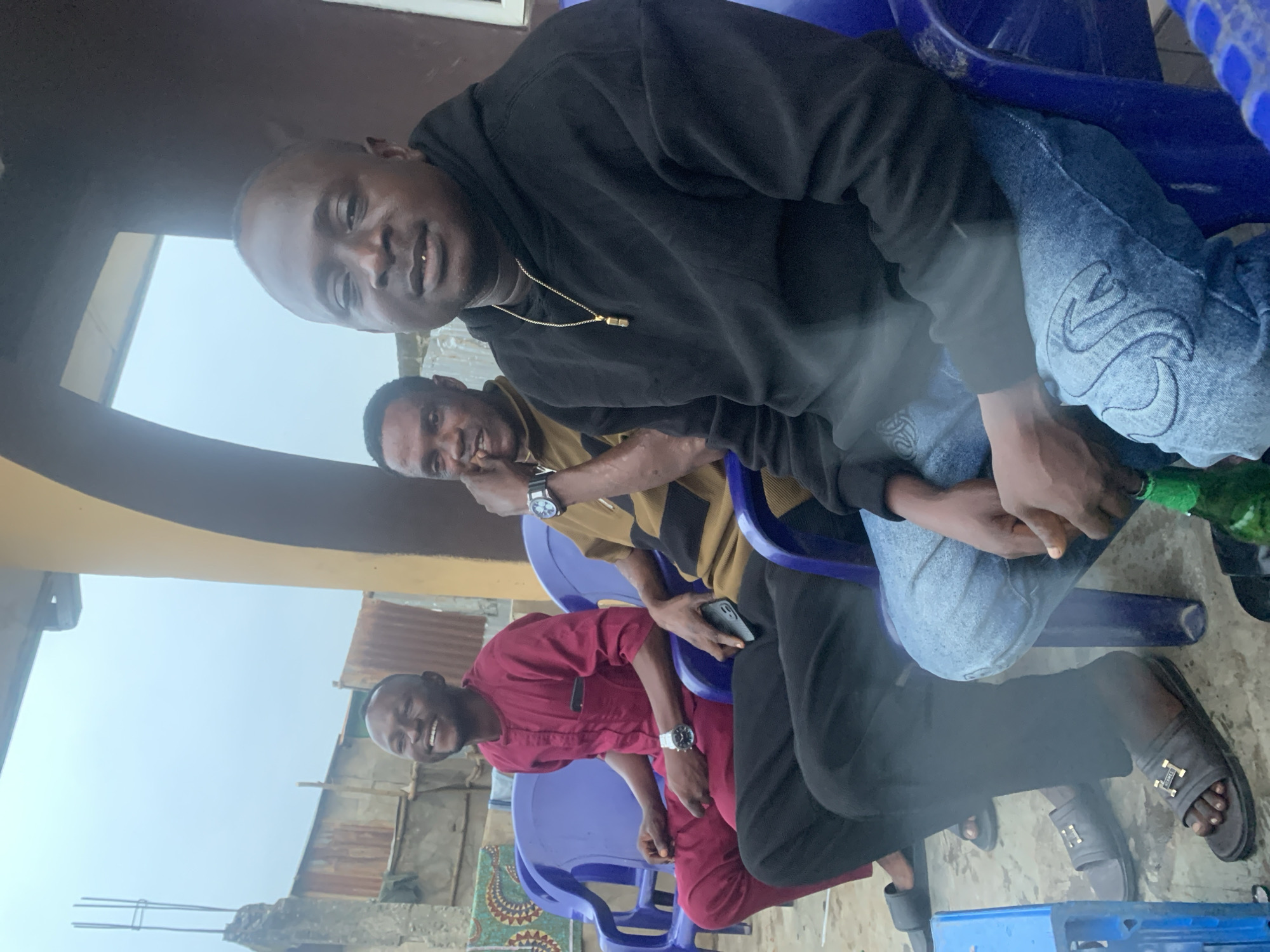 Three men are seated on blue plastic chairs outdoors, with a cloudy sky and a building structure in the background. The man on the left, wearing a maroon traditional outfit, is smiling and looking to his left. The man in the middle, in a brown and black striped shirt, is looking forward and resting his chin on his hand. The man on the right, wearing a black hoodie and jeans, is looking at the camera with a slight smile. There are a few objects visible, including cell phones in the hands of the middle and right man, a watch on the wrist of the man on the left, and a green bottle on the floor in front of the man on the right. The environment appears to be a simple outdoor setting, possibly a yard or courtyard. The overall impression is a casual gathering of friends. The scene is consistent with an outdoor space in Shagamu, Nigeria, given the architectural elements and the style of clothing.