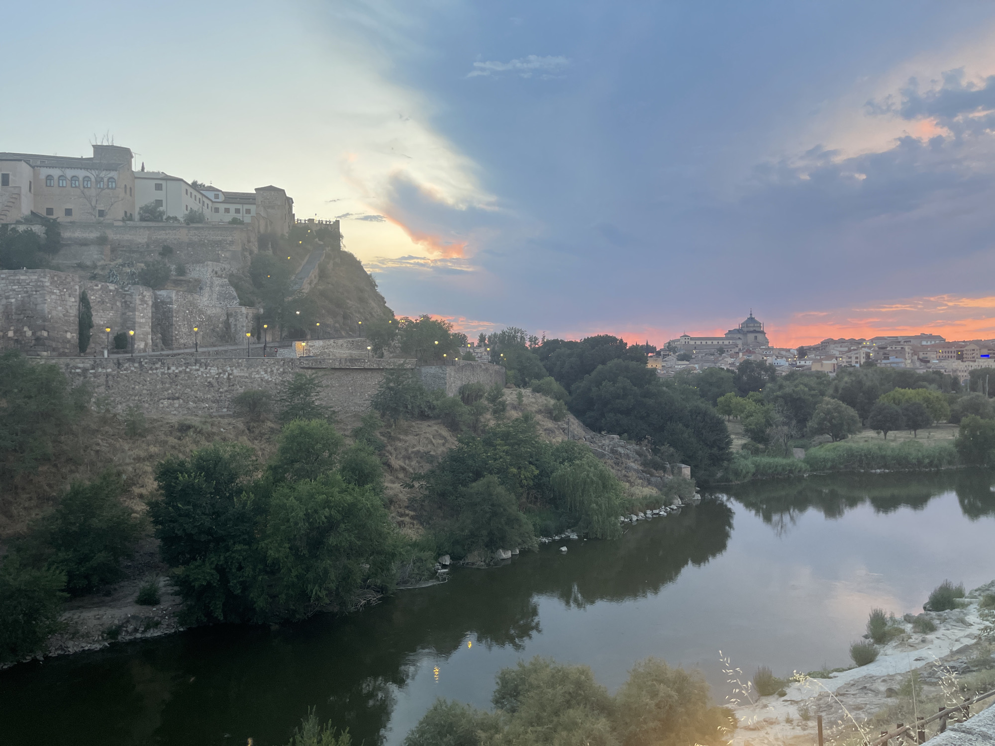 The image depicts a view of the city of Toledo, Spain, at dusk. In the foreground, a wide, calm river reflects the muted colors of the sky. The riverbanks are lined with lush green trees and vegetation. On the left side of the image, a steep hillside is covered with ancient stone fortifications and buildings, appearing to be part of the historic city walls and structures. Scattered streetlights are visible along pathways on the hillside. In the background, the Toledo skyline is visible, dominated by a large, domed cathedral, characteristic of Spanish cities. The sky displays a gradient of blues, purples, and pinks, indicating the setting sun, with some scattered clouds. The overall impression is one of historical grandeur and natural beauty during the twilight hours.