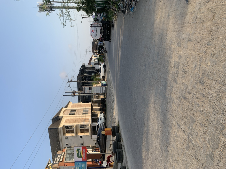 The image depicts a street scene in Ikota, Nigeria, under a clear, light blue sky with bright, even illumination suggesting daylight. The paved road, exhibiting some wear and tire marks, extends into the distance, narrowing slightly.
On the left side of the foreground, a stack of approximately ten black tires is present on a rough, unpaved shoulder. Adjacent to the tires, an orange barrier or drum is visible. A male individual, wearing a red long-sleeved shirt, dark trousers, and a white cap, is seated on a wooden bench or structure behind the tires, facing towards the road.
Further down the left side, a multi-story building with a dark roof and light-colored walls houses businesses, identifiable by signage including "THE VILLAGE BISTRO," "EXPRESS MALL," "La Monique," and "BIZEMARKET." Several vehicles are parked on the paved area in front of this building: a dark grey sedan, a white sedan, and a white SUV. A multi-panel signpost stands nearby.
Further down the street on the right side, a dark-colored multi-story building is visible, with a white SUV parked in front of it. On the right side of the road, a black flatbed truck with a large white tank-like container is parked. The tank displays a "G STRONG TOWER" logo and the phone number "070556644510." A male individual, dressed in a dark shirt and dark trousers, stands near the rear of this truck.
In the center of the road, a motorcycle with a single rider is in motion, heading away from the viewer. Utility poles with numerous overhead wires line both sides of the street. On the far right, a tall cellular communication tower is prominent. The right shoulder of the road appears unpaved and contains scattered debris and refuse. Sparse trees and vegetation are visible along the right side and in the background.