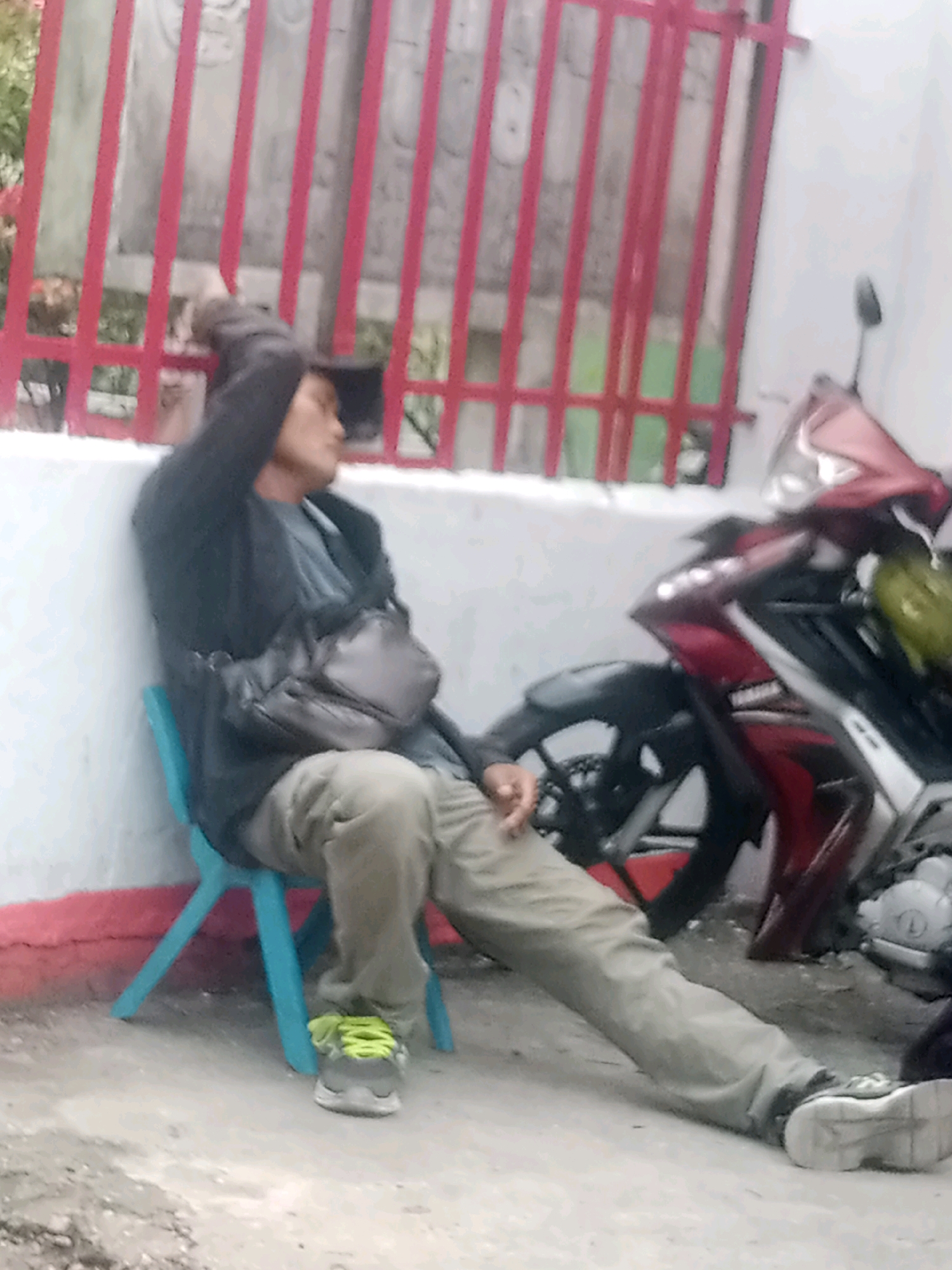 A person is seated on a blue plastic chair on a concrete surface next to a white wall. The person is wearing a dark jacket, a grey t-shirt, light-colored pants, and grey sneakers with neon green laces. A dark cross-body bag is slung across the person's chest. The person has their right hand raised to their head, and their eyes are closed or looking down. Behind the person, a red metal fence with vertical bars is visible, and beyond that, there appear to be some structures or vegetation. To the right of the person, a red and black motorcycle is parked. The lighting suggests it is daytime. The scene is consistent with an outdoor area in Pekanbaru, Indonesia, possibly a residential or commercial street.