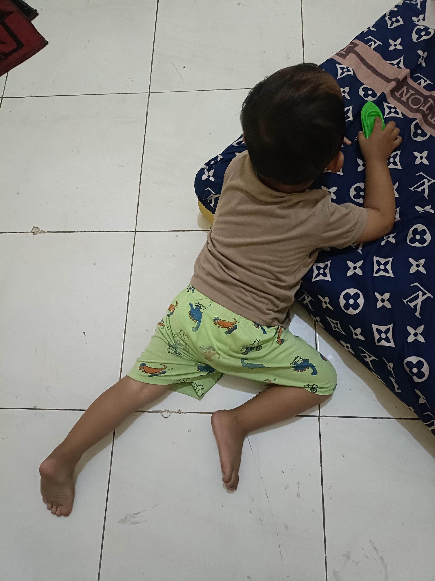 A young child is lying on a tiled floor, on their stomach, next to a patterned pillow. The child is wearing a tan t-shirt and light green shorts with a dinosaur pattern. The child is holding a small green object in their right hand, and their left leg is extended out to the side, with their foot pointing upwards. The floor is made of white tiles with dark grout lines. A dark red mat with a black border is visible in the upper left corner of the image. The patterned pillow is dark blue with a repeating pattern of white symbols. The scene appears to be indoors, possibly in a bedroom or playroom, given the presence of a child and a pillow. The image quality is average, with some slight blurriness. The lighting appears to be from overhead. There is no visible indication of the specific location within Kediri City, Indonesia, beyond the general assumption of an indoor domestic setting.