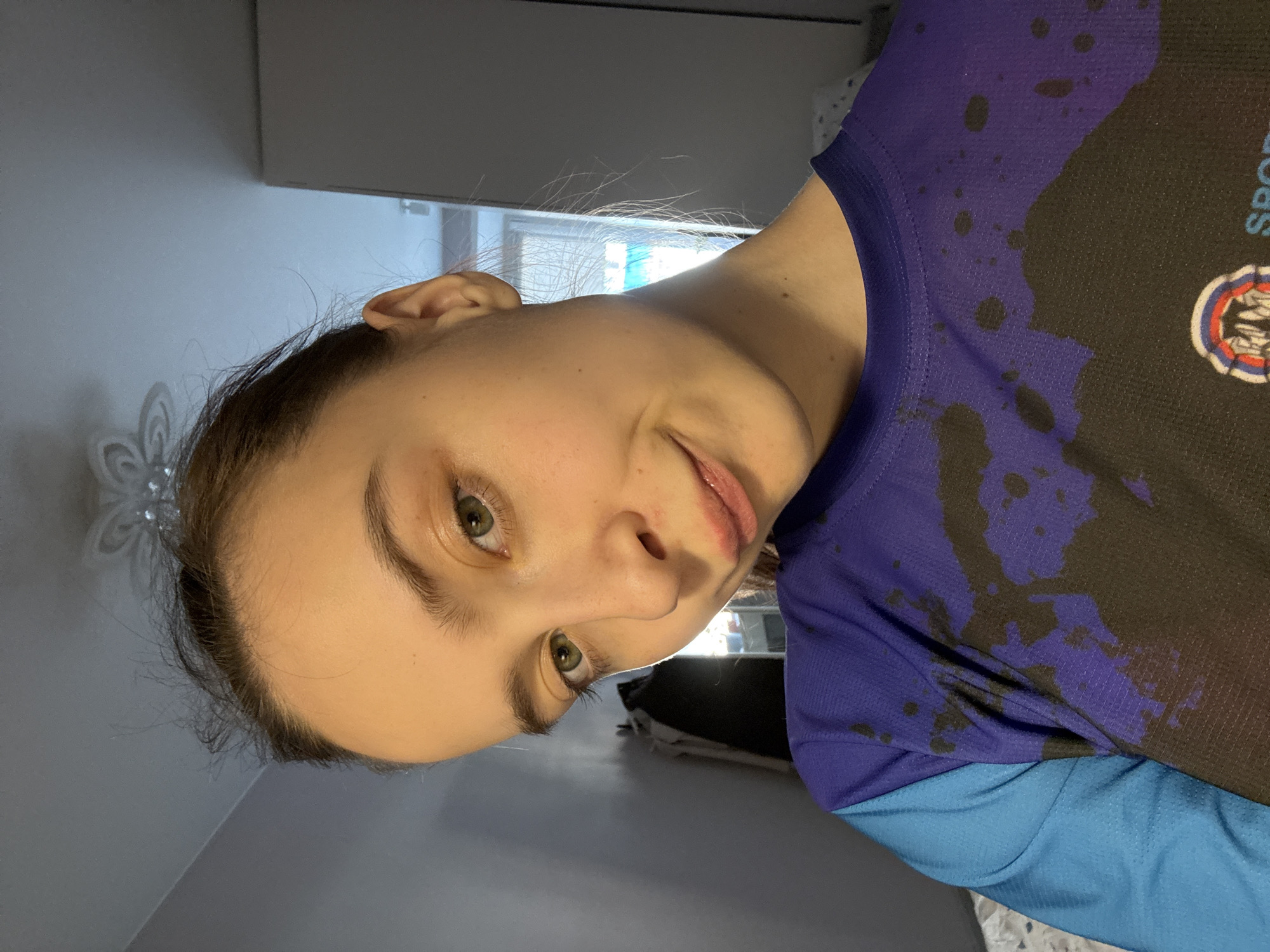 A young woman is depicted in a selfie. Her eyes are green. She is wearing a blue and black sports shirt with a team emblem. The location appears to be indoors, possibly in a residential setting with gray walls and furniture. The photo's orientation is slightly tilted. There is no definitive evidence to confirm the location is Chaikovsky, Russia.