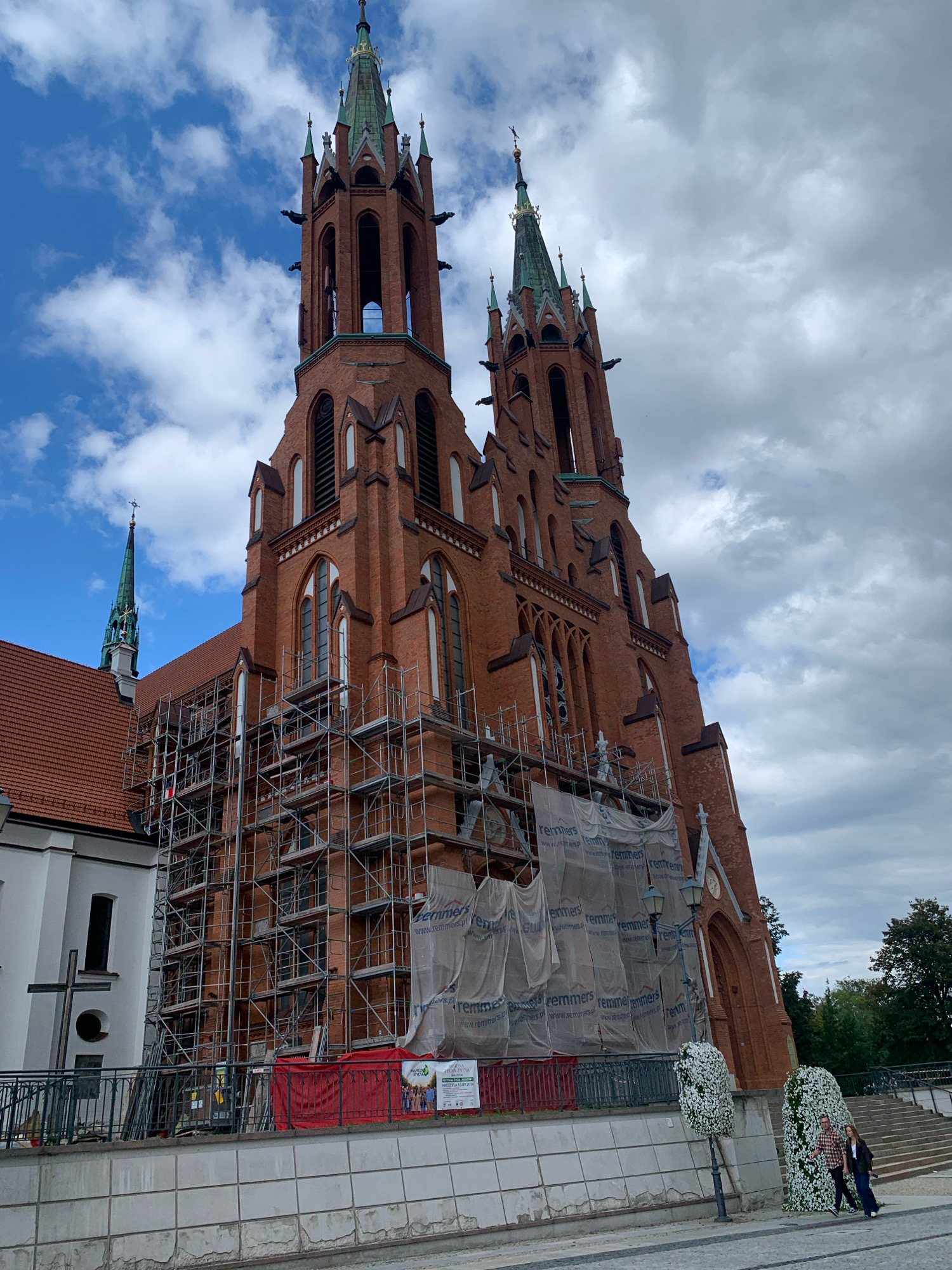 The image shows the exterior of the Basilica of the Assumption of the Blessed Virgin Mary in Białystok, Poland. The church is made of red brick and features two tall towers with green spires. Scaffolding covers a significant portion of the church's facade, suggesting ongoing renovations or construction. A white tarp, also branded with "remmers," is draped over some of the scaffolding. In the foreground, a sidewalk with a textured surface leads to the church. Two people, a man and a woman, are walking away from the camera on the right side of the frame. The sky is partly cloudy, with both blue sky and grey clouds visible. To the left of the main church structure, a white building with a red-tiled roof is visible, and a large metal cross stands in front of it. Flower decorations are placed near a lamppost and a large rectangular structure on the right side of the walkway.
