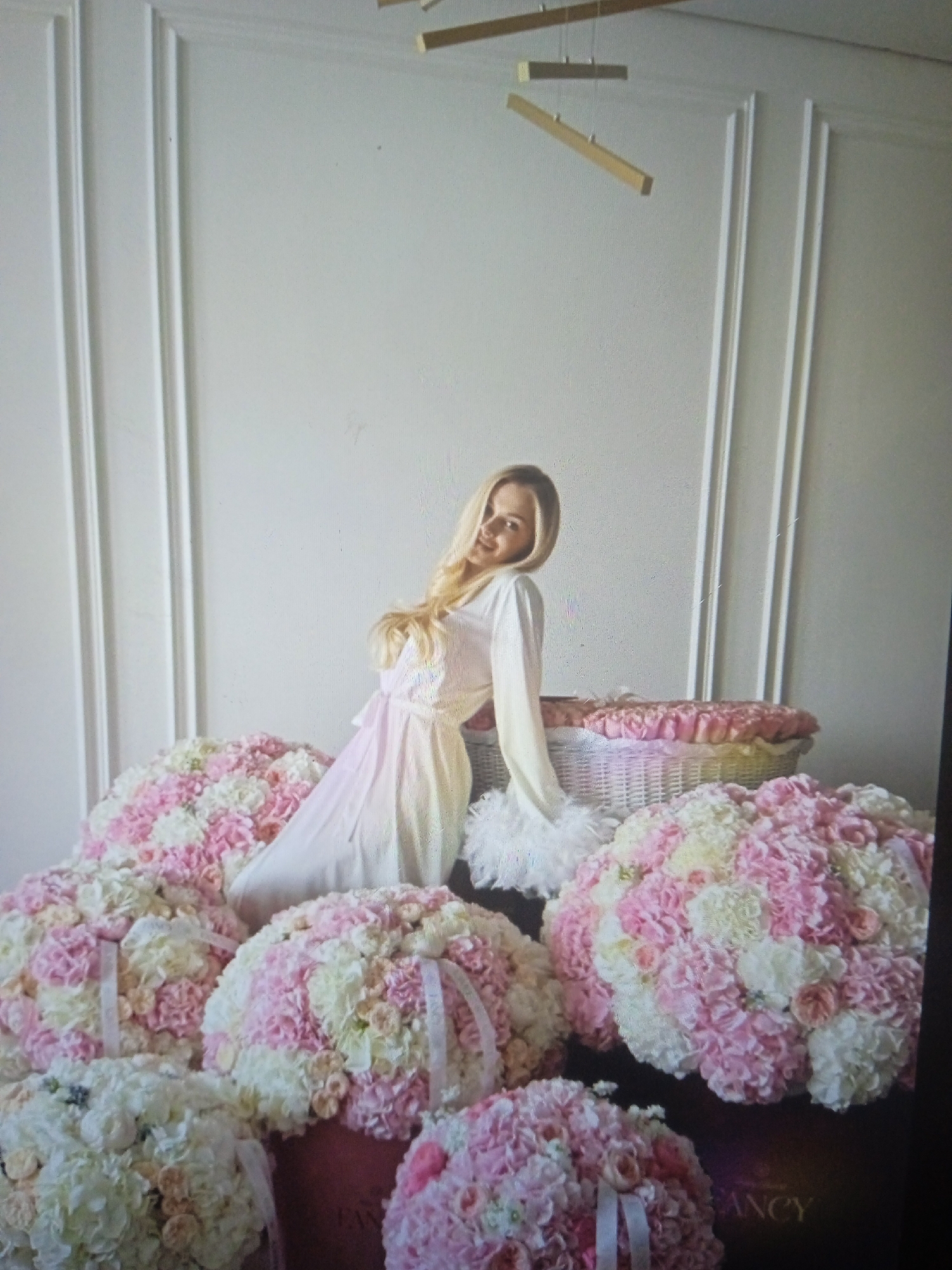 A blonde woman in a light pink dress and white feather trim is posing amidst a large display of pink and white floral arrangements. The woman is leaning on a wicker basket filled with similar flowers. The scene is set against a white wall with vertical decorative molding. The floor appears to be a dark color, and some of the floral arrangements are in brown containers with the word "FANCY" visible on one of them. The lighting suggests an indoor studio or staged environment. There is no visible indication of the city of Chernivtsi, Ukraine.