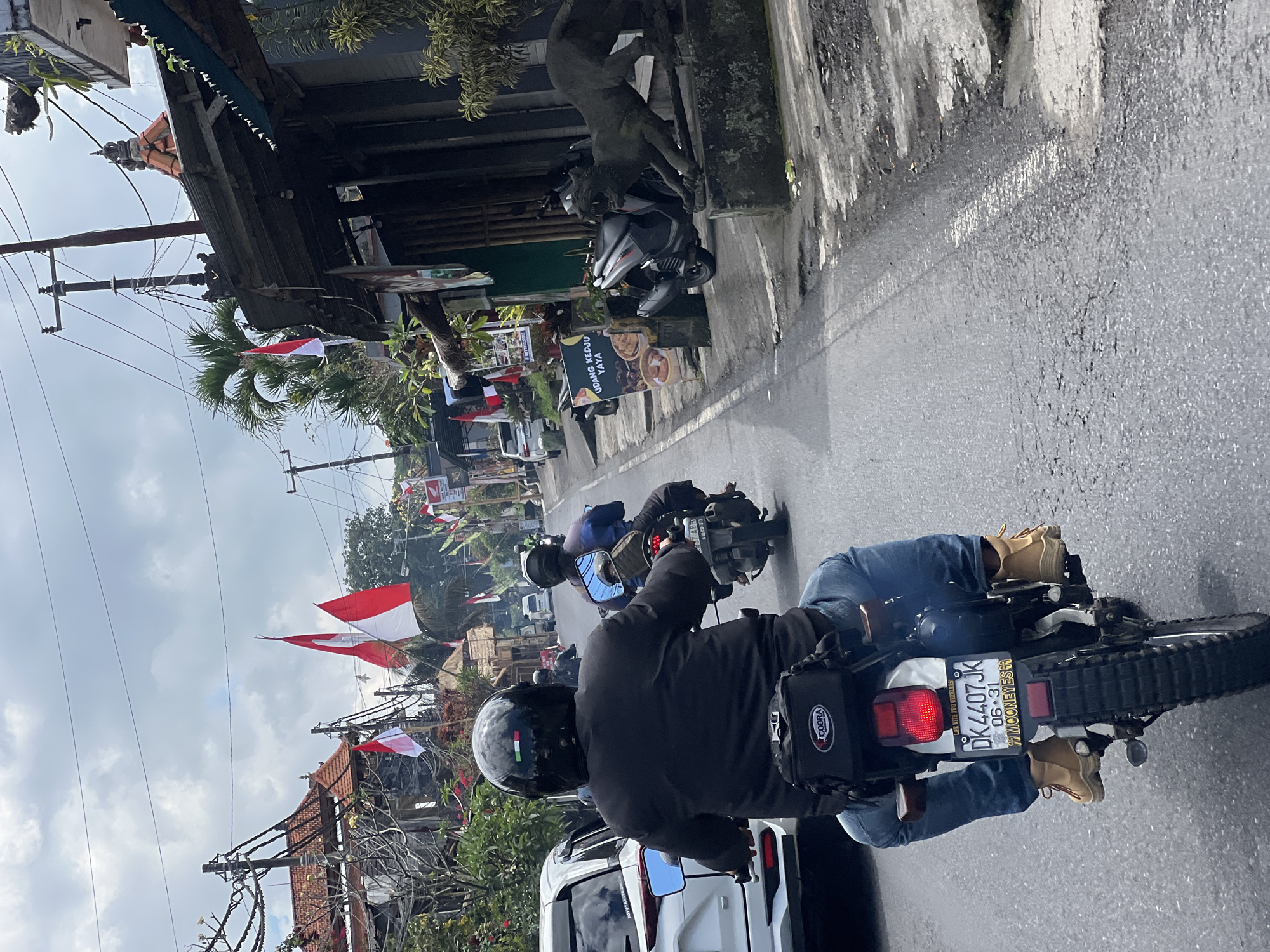An eye-level, medium shot captures a street scene in Payangan, Indonesia, dominated by a man riding a white motorcycle. He is viewed from behind, wearing a black helmet with an Italian flag decal, a black jacket, blue jeans, and tan boots. A black "COBRA" bag is attached to the rear of his motorcycle. The license plate reads "DK 4407JK 06-31 MOONEEYES". The road is paved and shows signs of wear. To the right of the road, there is a sidewalk with a concrete base supporting a dark grey sculpture of a crouching animal, possibly a panther, with its mouth open. Behind the sculpture is a building with a dark roof and a black sign. Several Indonesian flags, red and white, are visible on poles along the street, suggesting a national holiday or celebration. Other motorcycles and a white car are also present on the road. The sky is overcast with white clouds. The buildings lining the street appear to be shops or residences, with various architectural styles and some greenery visible.