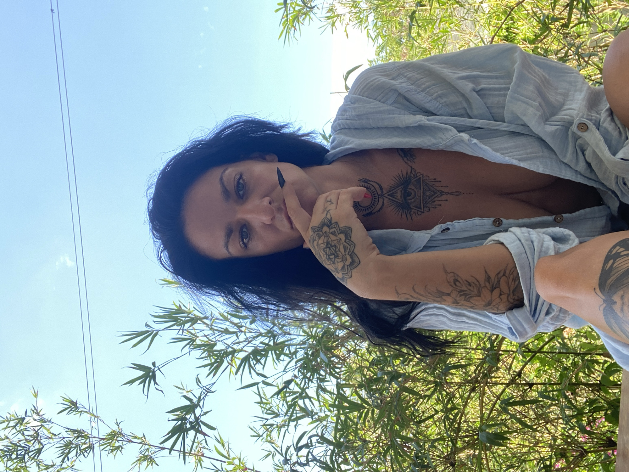 A low-angle, rotated, outdoor shot depicts a woman with dark hair and tattoos. She is wearing a light blue button-down shirt, partially unbuttoned. The woman is gazing directly at the camera. Her right hand is near her face, with her index finger touching her chin. Tattoos are visible on her chest, arms, and possibly legs. The backdrop features green foliage and a blue sky. Two thin wires are visible in the top left corner of the image. The location appears to be Mũi Né, Vietnam, based on the presence of tropical vegetation.