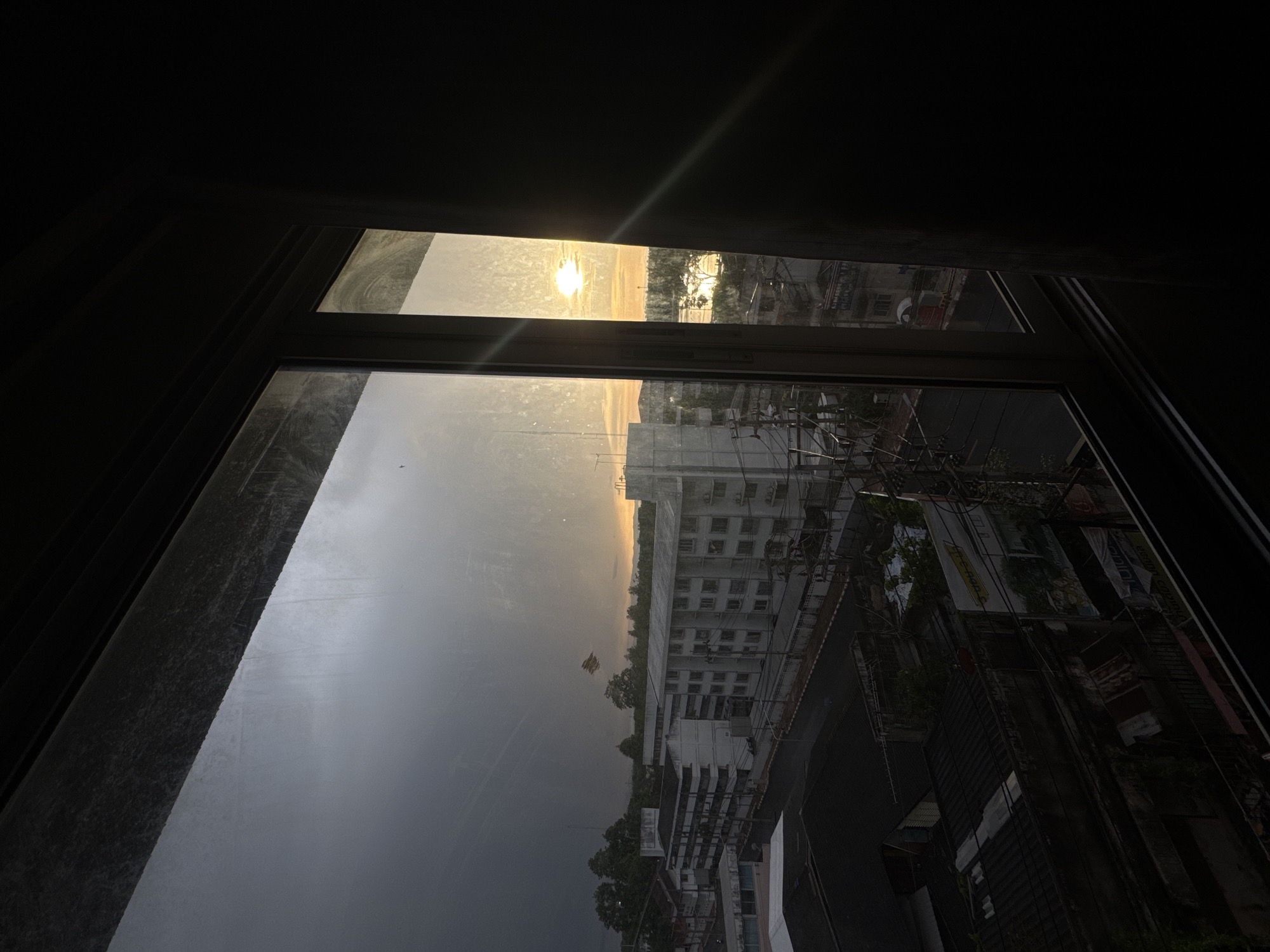 This image captures a view through a dirty window of a residential area in Surat Thani City Municipality, Thailand, at sunset. The sky is a dramatic mix of dark, stormy clouds on the left and a bright, sunlit area on the right, with the sun appearing as a hazy orb near the horizon. The cityscape below includes several multi-story apartment buildings with white facades and dark window frames, as well as lower-rise buildings with dark roofs. Electrical wires and poles are visible throughout the scene. The foreground is dominated by the dark interior of the room from which the photo is taken, with the window frame creating a strong border. There are no people or discernible animals visible in the image.