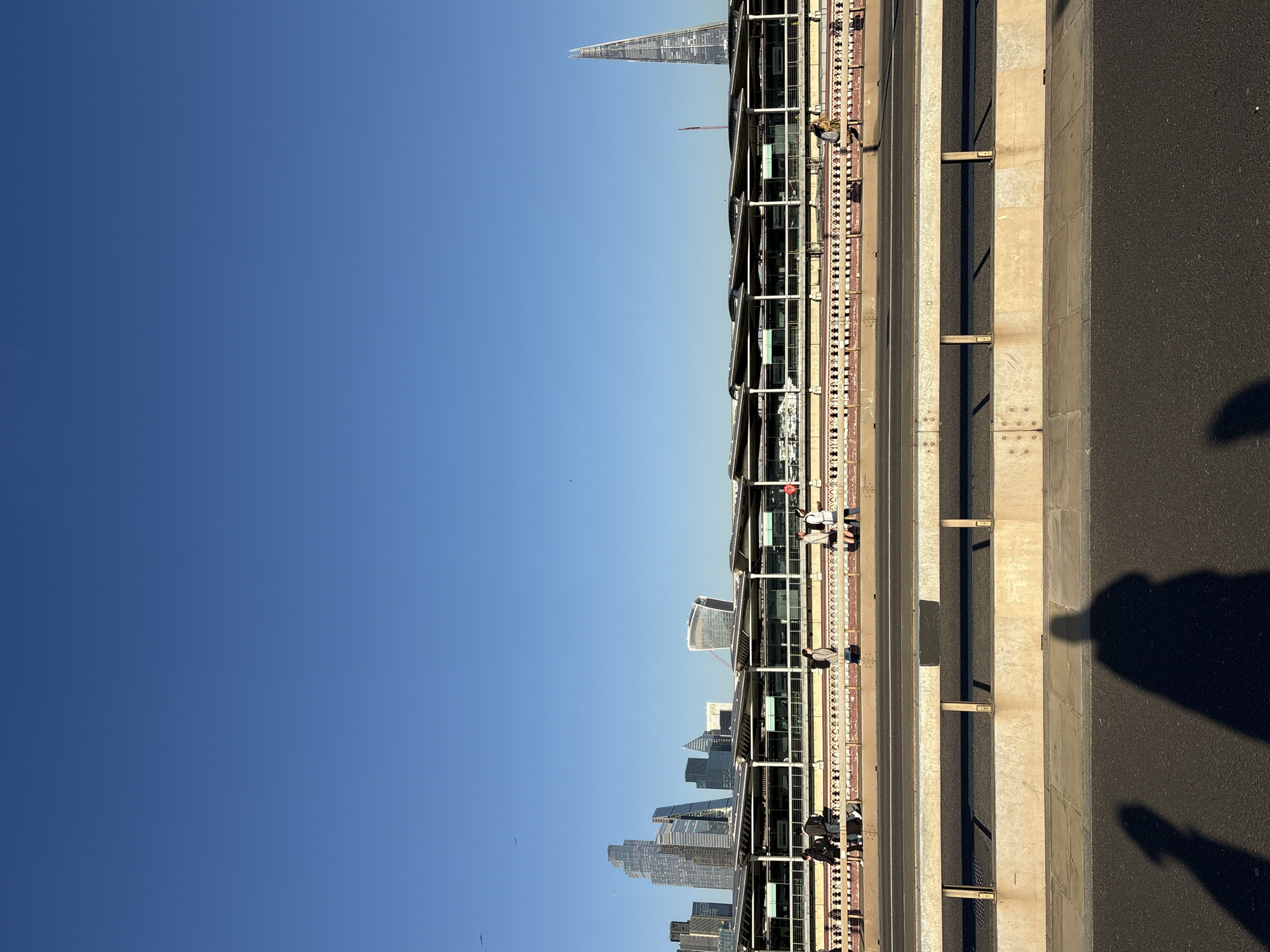 The image shows a view from Tower Bridge in London, United Kingdom, looking towards the city. The Shard is visible in the distance. A walkway along the bridge is visible, with people walking. The bridge structure, including railings and supports, is in the foreground, and a shadow of someone is also visible in the foreground. The sky is clear and blue.