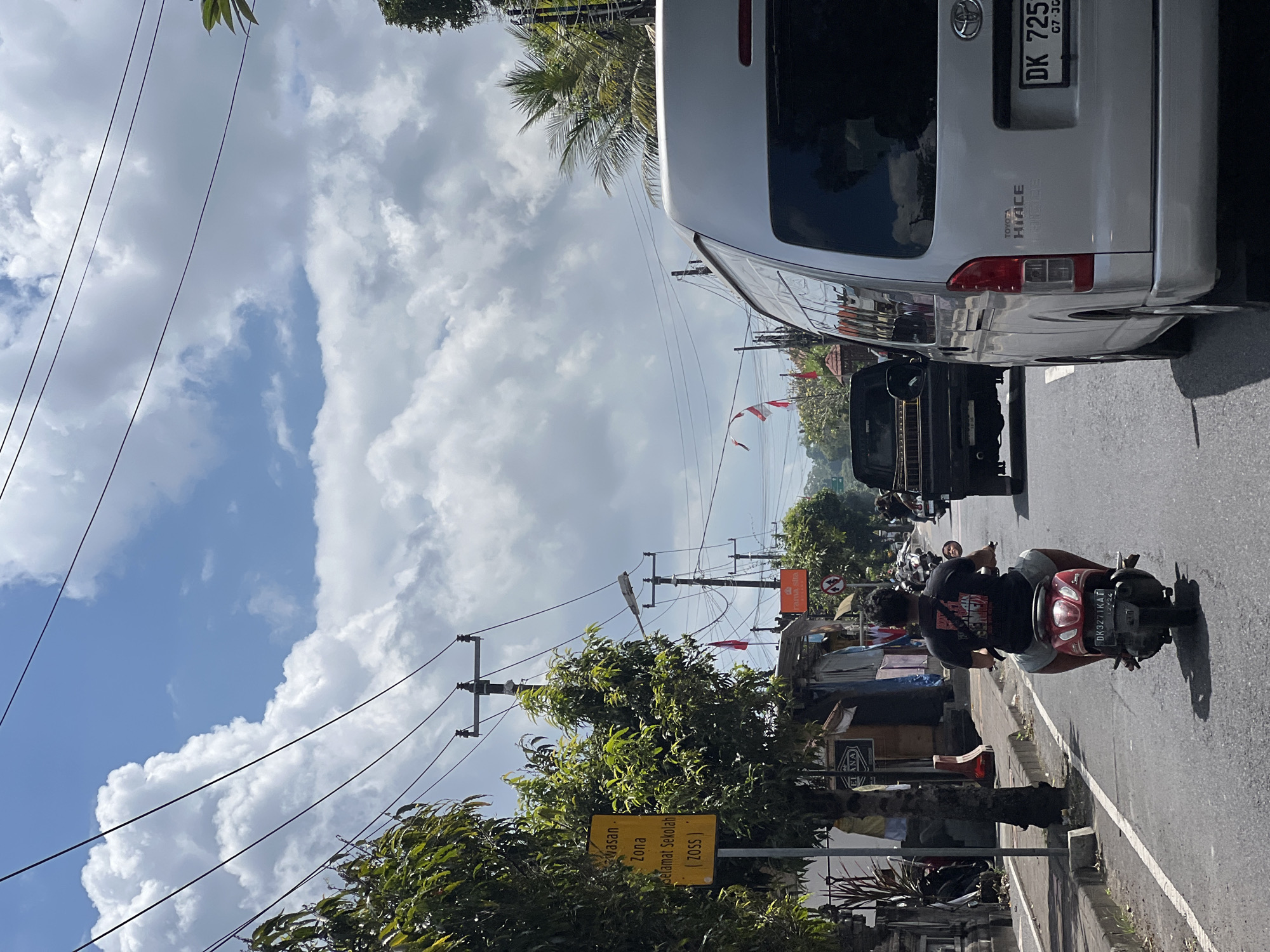 The image captures a street scene in Payangan, Indonesia, under a bright, cloudy sky. A silver Toyota Hiace van dominates the foreground, viewed from the rear right, revealing its taillight and license plate, "DK 7257 JC". To the left of the van, a black pickup truck is visible. Further ahead, a person is riding a red scooter in the same direction as the van, with their back to the camera. The scooter's license plate is "DK 3271 KA".
The road is paved and appears to be a typical street in a populated area, with trees lining the left side. A yellow sign is posted on the left, with Indonesian text including "ZONA" and "SEKITAR SEKOLAH". Utility poles with numerous wires are scattered throughout the scene, some with streetlights attached. Red and white Indonesian flags are seen fluttering in the distance. The buildings along the street are modest, with some appearing to be shops or small residences. The overall atmosphere suggests a sunny day with scattered clouds, characteristic of a tropical climate.
