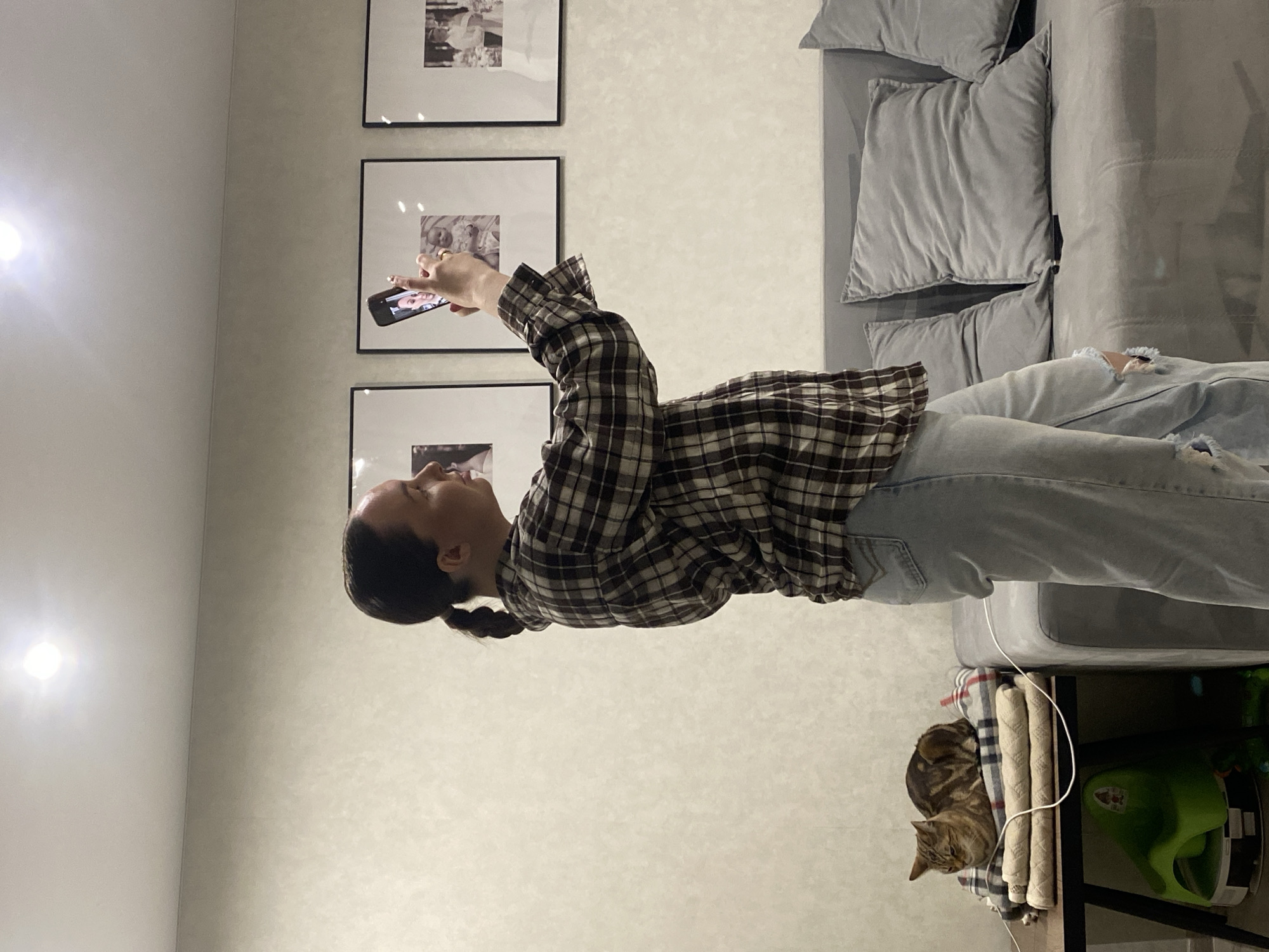 The image depicts an indoor scene, likely a living room, in Chaikovsky, Russia. The main subject is a woman, viewed from the waist up, facing right. She has dark hair styled in a braid and is wearing a brown and white plaid long-sleeved shirt paired with light blue ripped jeans. She holds a smartphone in her hands, its screen displaying an image of a person, possibly a child.
In the background, a grey sofa with several plump grey cushions occupies the right side of the frame. The wall behind the woman is a light, textured color, adorned with three framed pictures arranged vertically. These frames contain what appear to be black and white photographs, potentially depicting babies or children. Two bright spotlights are visible on the left side of the wall, illuminating the area.
In the lower right corner of the image, a striped tabby cat rests comfortably on a stack of folded items, which are placed on a small dark side table or shelf. A white cable, possibly a phone charger, snakes from the area near the sofa towards the items where the cat is resting. A green object, perhaps a pet toy or bowl, is partially visible underneath the side table. The lighting suggests an indoor setting, possibly during the evening or night. The woman's expression is neutral, and the cat appears relaxed. No legible text is visible in the image.