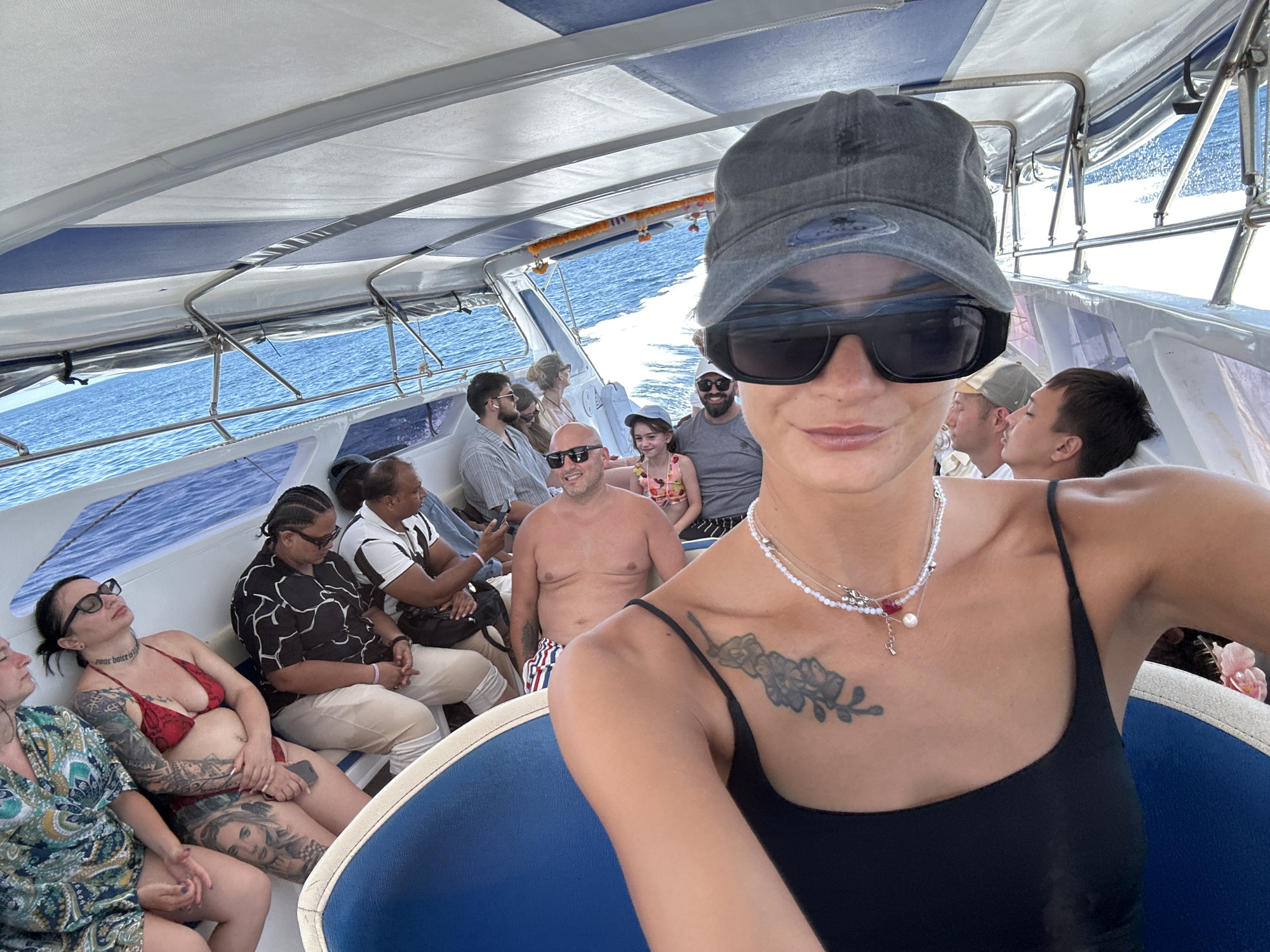 In Phru Nai, Thailand, a group of people are on a boat under a white and blue striped canopy. The water is blue and choppy. In the foreground, a woman wearing a black tank top, sunglasses, and a gray baseball cap is taking a selfie. She has a tattoo on her chest and is wearing a necklace. Behind her, other passengers are seated on the boat. To the left, a woman in a red bikini and another woman in a patterned dress are visible. Further back, several men and a young girl are also seated. The overall atmosphere appears to be a casual boat trip.