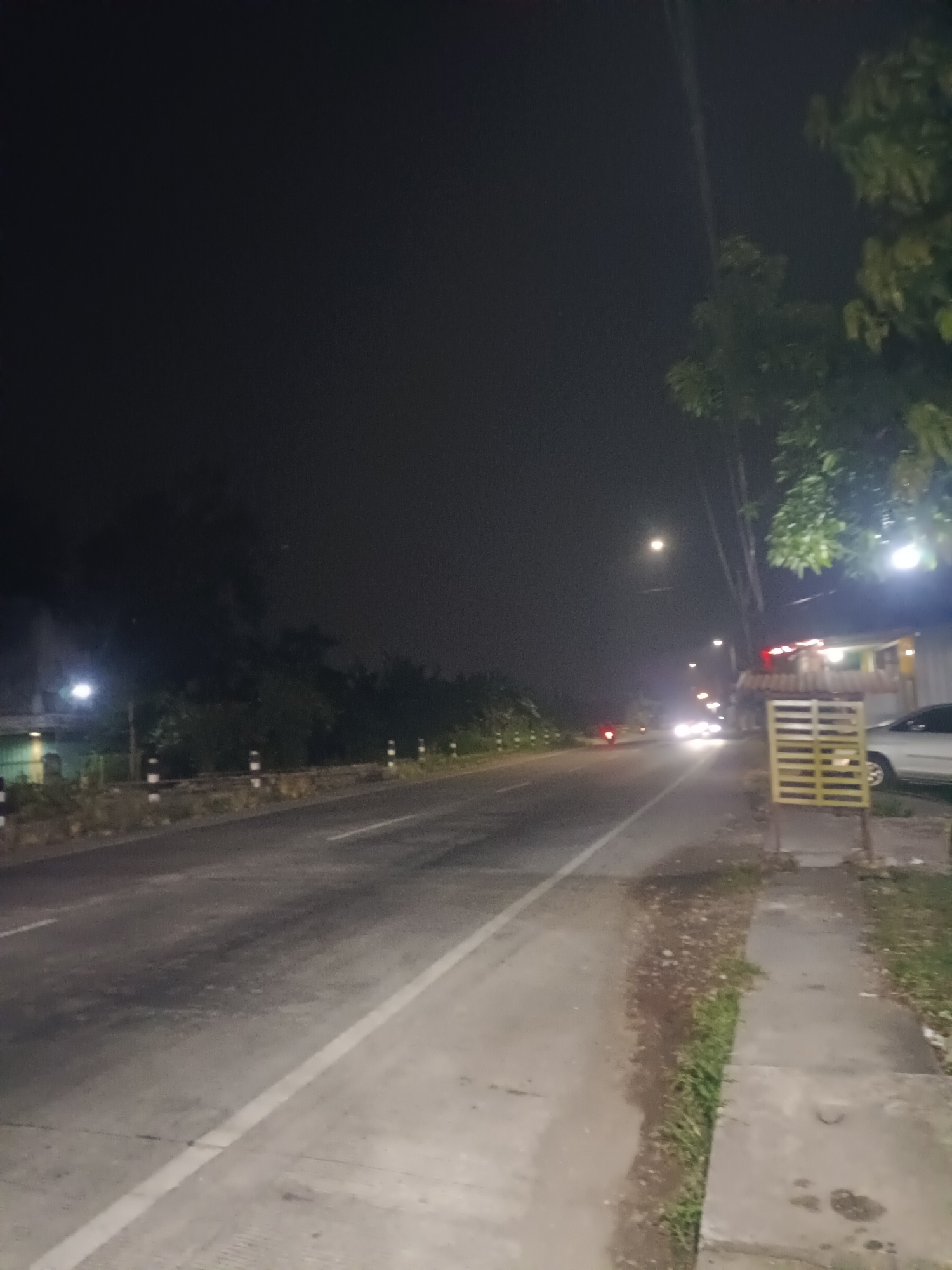 The image depicts a street scene at night in Kajeksan, Indonesia. A two-lane road stretches into the distance, illuminated by streetlights. Along the road's edge is a sidewalk with vegetation growing between it and the road. A parked silver car is visible adjacent to a building with a yellow metal structure. Traffic includes a motorcycle and a car with its headlights on in the distance. The sky is dark, and trees line the background.