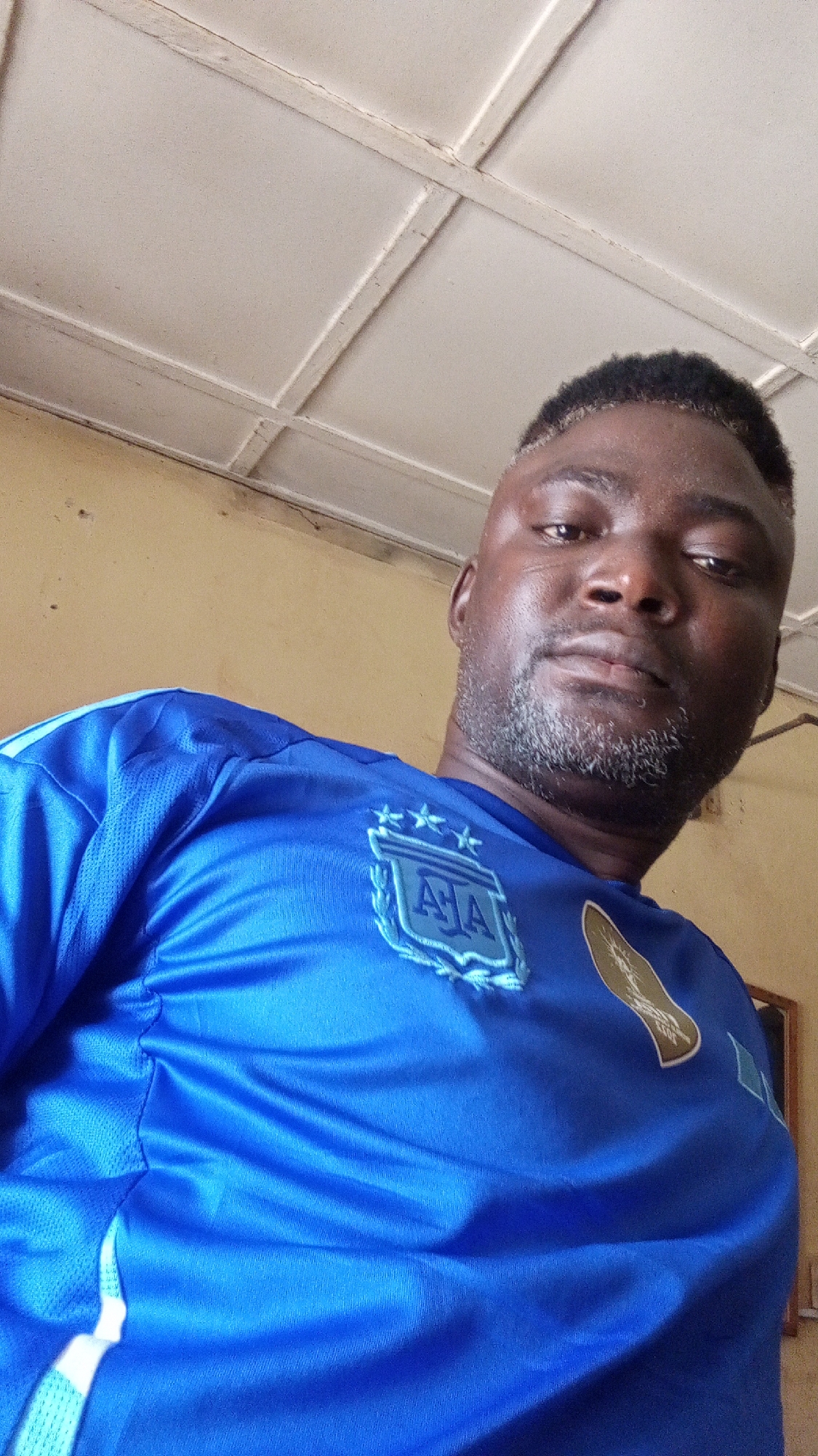A medium shot captures a middle-aged man wearing a blue jersey with Argentina's insignia on the left breast. He appears to be indoors, with a white, paneled ceiling visible in the background. The wall is painted a pale yellow. The location is presumed to be Sabon Tasha, Nigeria.
