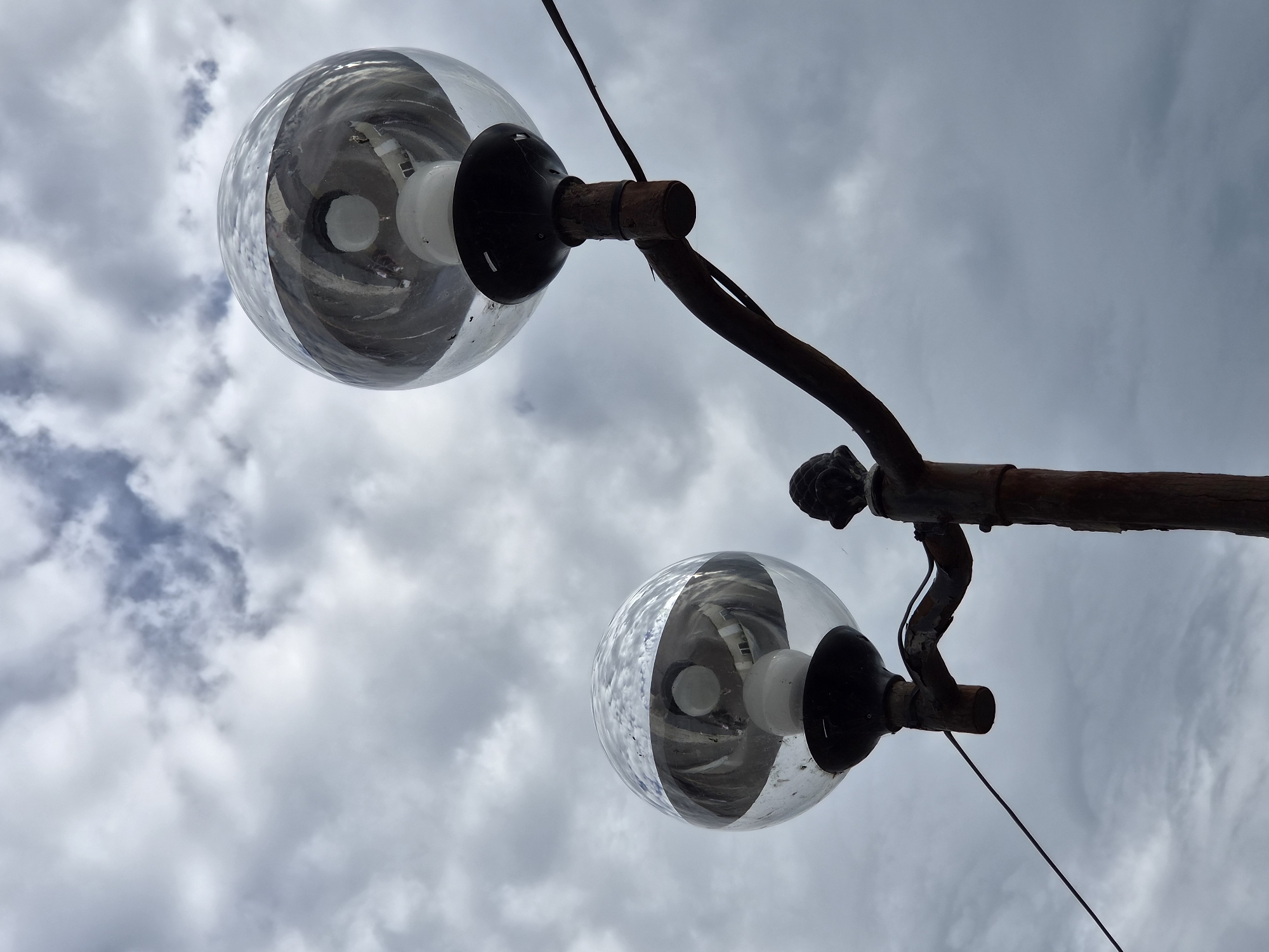 Two spherical glass street lamps are mounted on a rusty metal support. The background is a cloudy sky. It appears to be in the city of Sochi, Russia.
