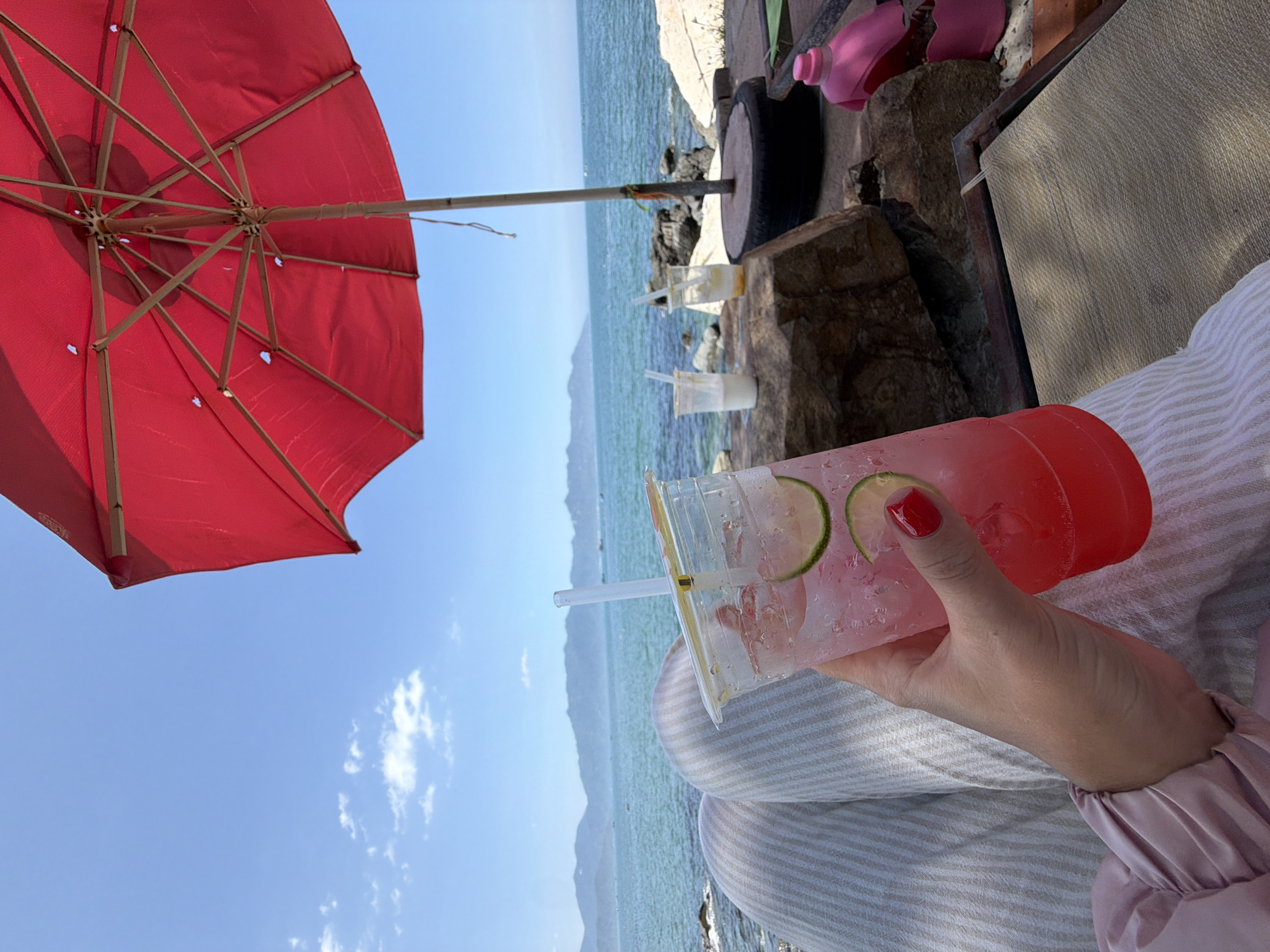 The image displays a coastal scene in Vĩnh Lương, Vietnam, under a clear blue sky with scattered white clouds. A person's right hand, with red nail polish, holds a clear plastic cup containing a pink beverage with ice cubes and two lime slices; condensation is visible on the cup's exterior. The person is wearing light-colored, vertically striped pants and a light pink long-sleeved top with ruched cuffs. A red beach umbrella, with a visible wooden pole anchored in a black tire-like base, occupies the upper right side of the frame, casting shade. The turquoise sea extends from the mid-ground to a distant blue mountainous horizon. On a large dark rock in the mid-ground, two other partially consumed clear plastic cups with straws are positioned. The immediate foreground includes additional large rocks, a section of a light-colored woven sun lounger, and a pink plastic bottle.