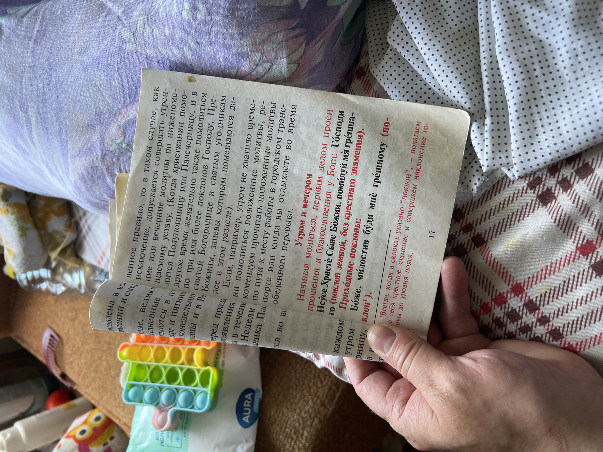 The image displays a close-up, overhead view of an open book with Cyrillic text, resting on a patterned fabric. The book's pages are slightly yellowed with age. To the left of the book, a colorful pop-it toy is visible, as well as part of a package labeled "AURA". A hand, with visible fingernails, is holding the book open. The background consists of a light-colored, patterned fabric with a floral or abstract design in shades of purple and white. The scene appears to be indoors, possibly in a domestic setting. There are no specific landmarks or details that would indicate the location of Кемуль, Russia.