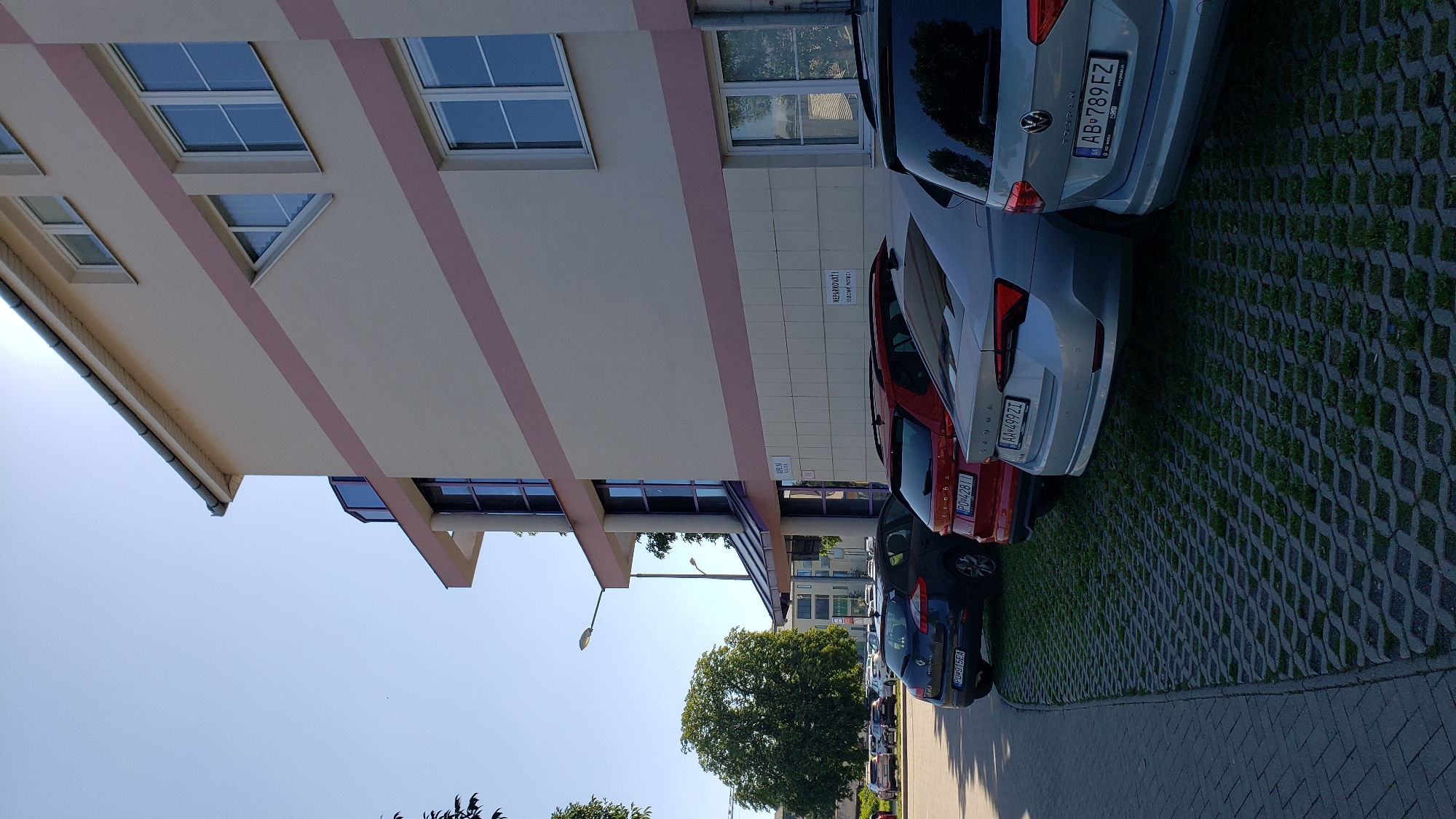 The image shows a modern, light-colored building with pink horizontal stripes, likely an office building or residential complex, located in Prešov, Slovakia. In front of the building, a row of cars is parked on a patterned concrete surface with grass growing through it, adjacent to a paved walkway. The cars are diverse, including a dark blue hatchback, a red SUV, a silver sedan, and a light gray SUV. The sky is clear and blue, suggesting a sunny day. A street lamp is visible on the left side of the image, along with a large green tree. A sign on the building reads "NEPARKOVAŤ!" which means "DO NOT PARK!" in Slovak, indicating parking restrictions. The overall scene is an outdoor urban environment on a clear day.