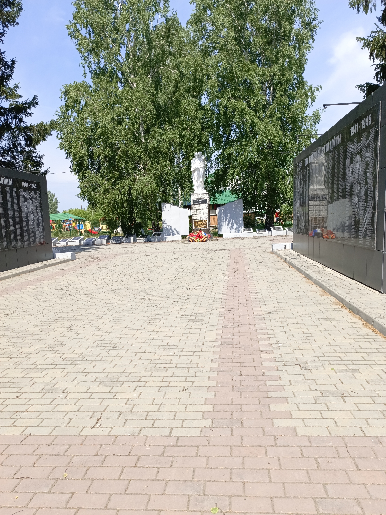 This is a ground-level, wide-angle photograph taken outdoors in Zalesovo, Russia, on a sunny day. The foreground is dominated by a paved walkway made of light tan and pink rectangular bricks laid in a repeating pattern. A path of pink bricks runs down the center of the walkway, extending into the middle ground.
In the middle ground, on the left side of the image, is a dark, multi-panel monument. The panels appear to be made of polished black stone, inscribed with rows of names and dates, with one panel showing a bas-relief image of figures. The inscription "1941-1945" is visible on one of the panels. To the left of this monument, a dark green coniferous tree is partially visible. In front of the monument, a white statue of two figures stands on a pedestal. A wreath of red flowers is placed at the base of the statue. Further back, a white, rectangular structure with a green roof is visible. Beyond that, a colorful playground with slides and swings can be seen.
On the right side of the image, a large, dark, multi-panel monument similar to the one on the left is present. It is also made of polished black stone with inscribed names and dates, and bas-relief images. The inscription "1941-1945" is clearly visible. Red floral wreaths are placed at the base of this monument as well.
The background features a large number of mature deciduous trees with dense green foliage, suggesting it is summertime. The sky is blue with some wispy white clouds. The overall scene depicts a war memorial with a surrounding park-like environment.