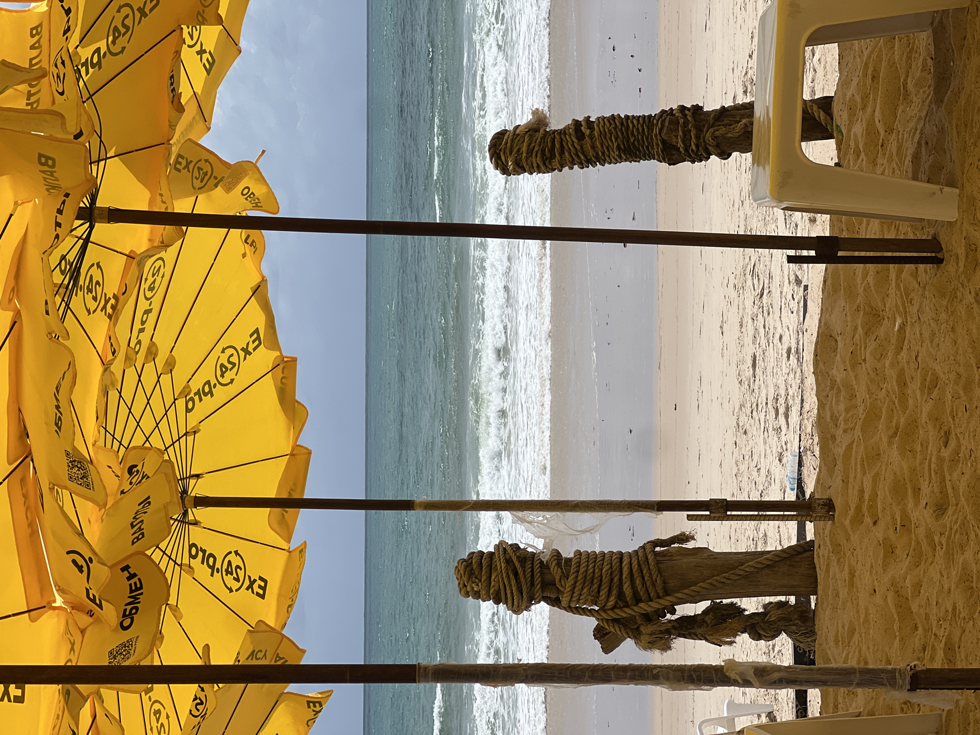 The image shows a beach scene with a yellow beach umbrella and a white plastic chair. The scene is viewed from under the umbrella, looking out towards the ocean. The beach is sandy, and the ocean has gentle waves. The scene takes place at an unspecified location.