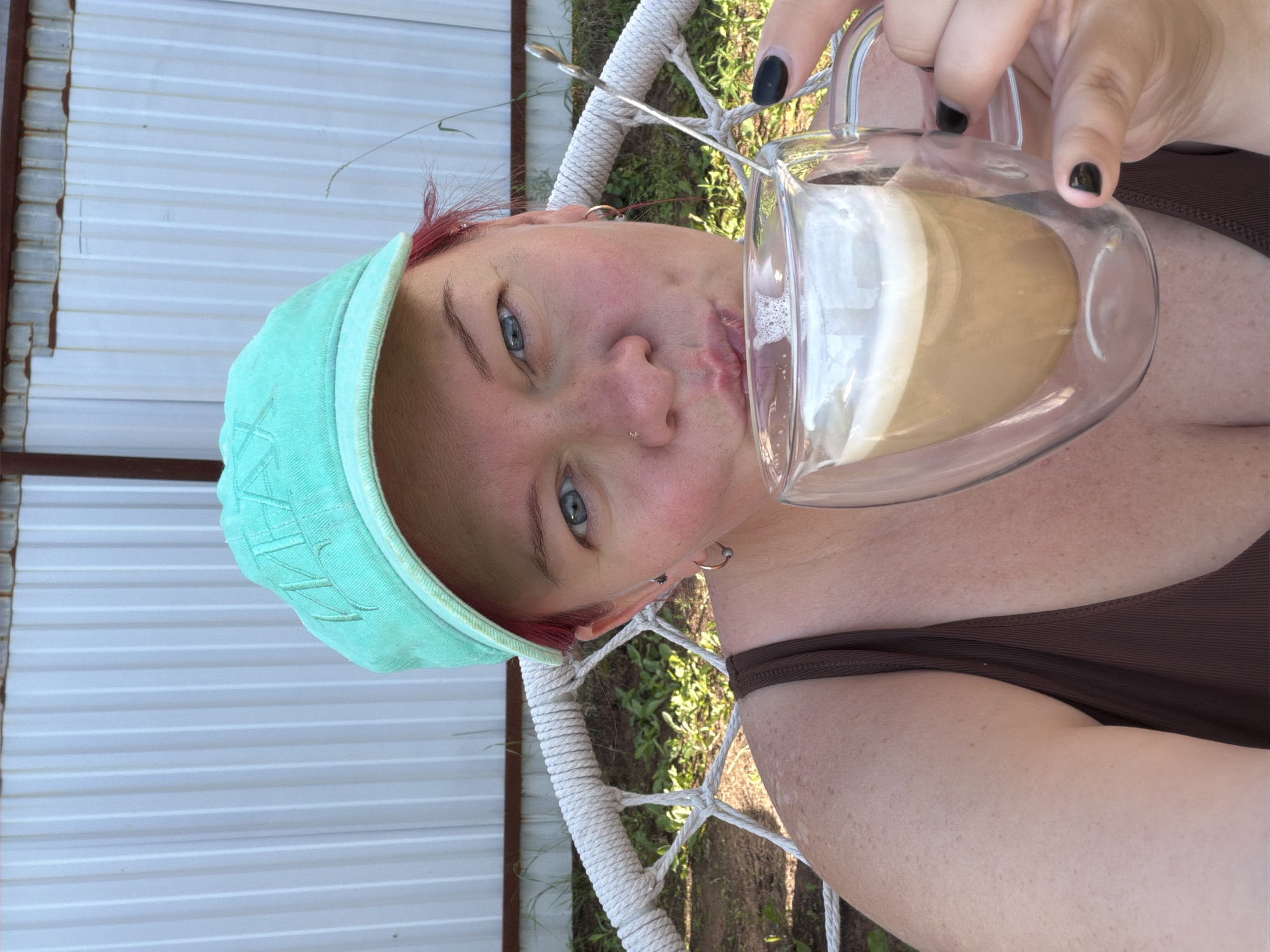 A woman wearing a teal baseball cap and a brown tank top sits in a white rope hammock chair, drinking from a clear double-walled mug filled with a latte. She has blue eyes, red hair, and a nose piercing. Her left hand, with black nail polish, holds the mug, and her right arm is visible on the side. The background shows a white corrugated metal fence and green foliage. The scene appears to be outdoors, possibly in a backyard or recreational area in Chaikovsky, Russia.