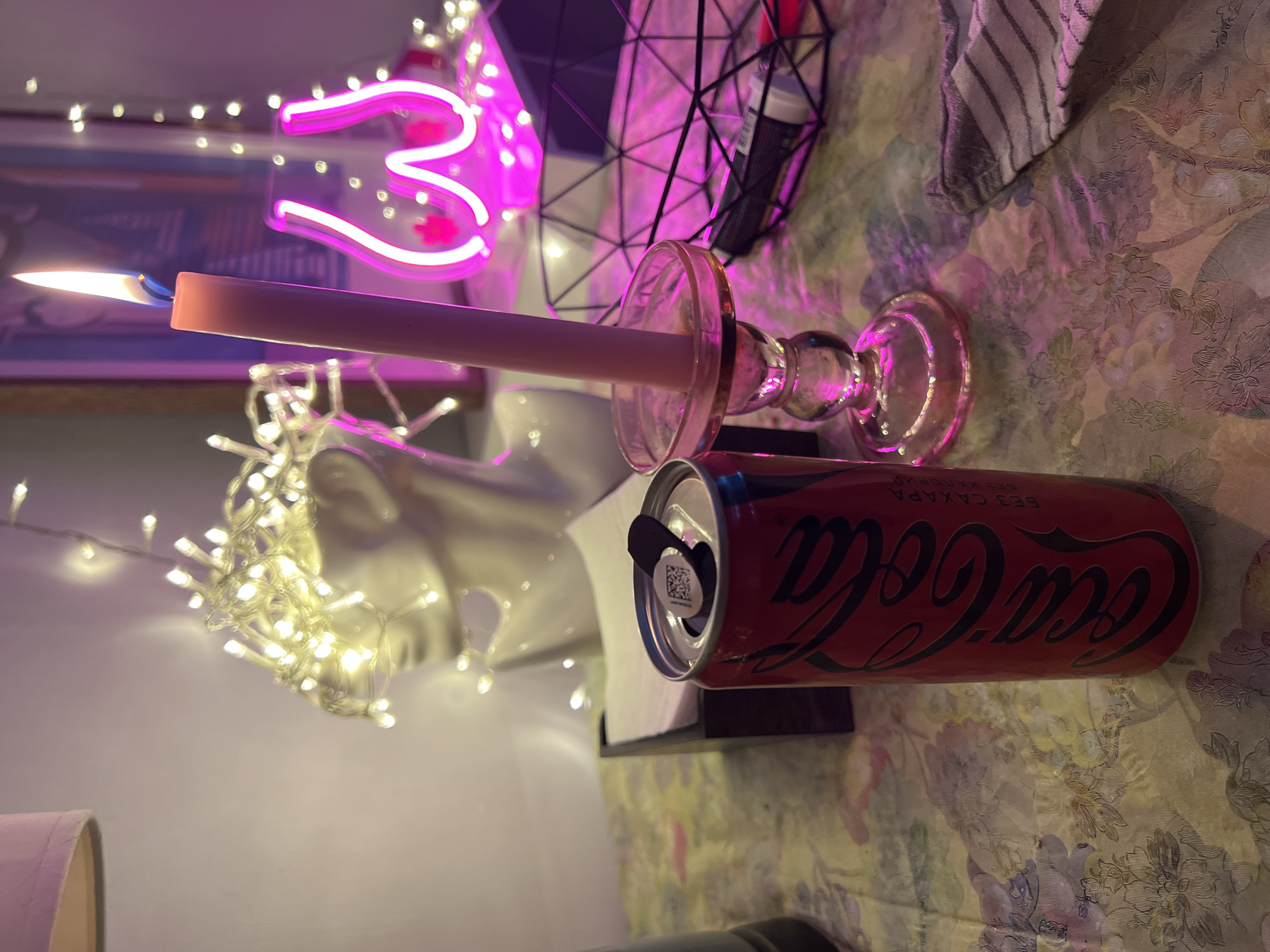 The image shows a table with a Coca-Cola can, a burning candle in a glass holder, a napkin holder, and a wire basket. In the background, there is a bust decorated with string lights, a framed picture, and a pink neon sign shaped like breasts. The table is covered with a floral patterned cloth, and the lighting is a mix of warm yellow from the candle and string lights, and pink from the neon sign. The scene appears to be set indoors, possibly in a residential or retail space. The location in Khimki, Russia, cannot be determined from the image.