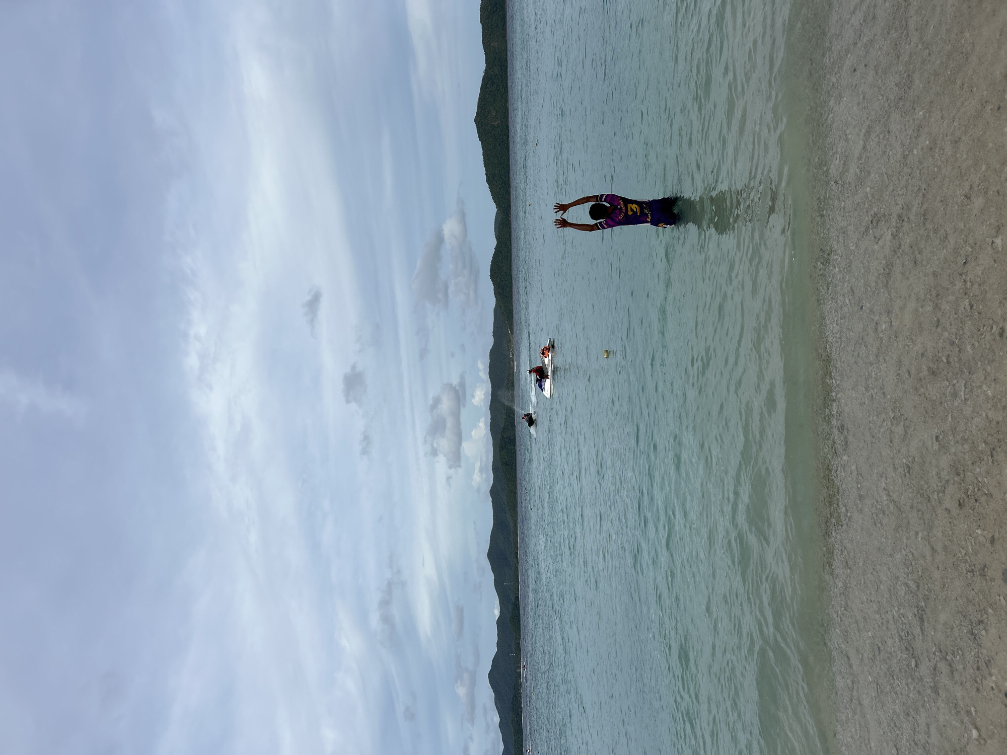 This is a photograph taken at Phru Nai, Thailand, depicting a serene coastal scene. In the foreground, a person stands waist-deep in the clear, shallow water, facing away from the camera with their arms raised. They are wearing a purple and yellow shirt with the number '3' visible on the back and dark shorts. The water is calm, with gentle ripples breaking on the sandy, pebble-strewn shore.
In the middle ground, several jet skis are active on the turquoise water. One jet ski is closer to the viewer on the right, with a rider visible. Further out, two other jet skis can be seen. A small yellow buoy floats in the water near the center of the frame.
The background features a chain of lush, green islands, their slopes covered in dense vegetation. The sky is overcast with a pattern of light gray and white clouds, suggesting a possibly humid or impending weather event. The overall impression is one of a recreational day at the beach with water sports in progress.