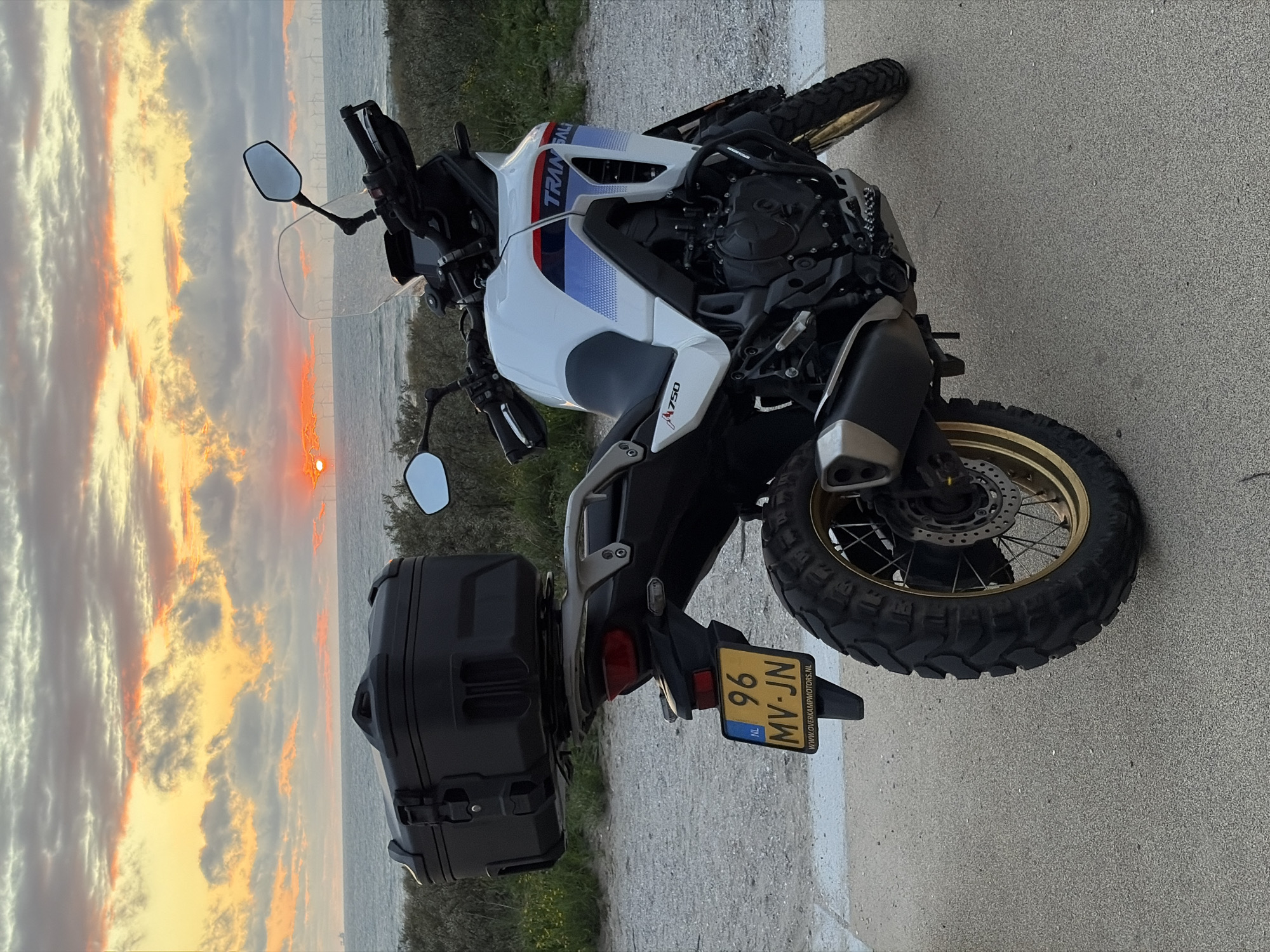 A white and blue Honda Transalp motorcycle with a black top case is parked on a paved surface next to a grassy area. The motorcycle has a license plate reading "96 MV-JN" and a website "WWW.OVERKAMPMOTORS.NL". In the background, a body of water, possibly the sea, is visible with windmills in the distance. The sun is setting over the water, casting a warm glow on the clouds. The scene appears to be a coastal area, possibly Scheveningen in The Hague, Netherlands, given the windmills and coastal landscape. There are no people visible in the image.