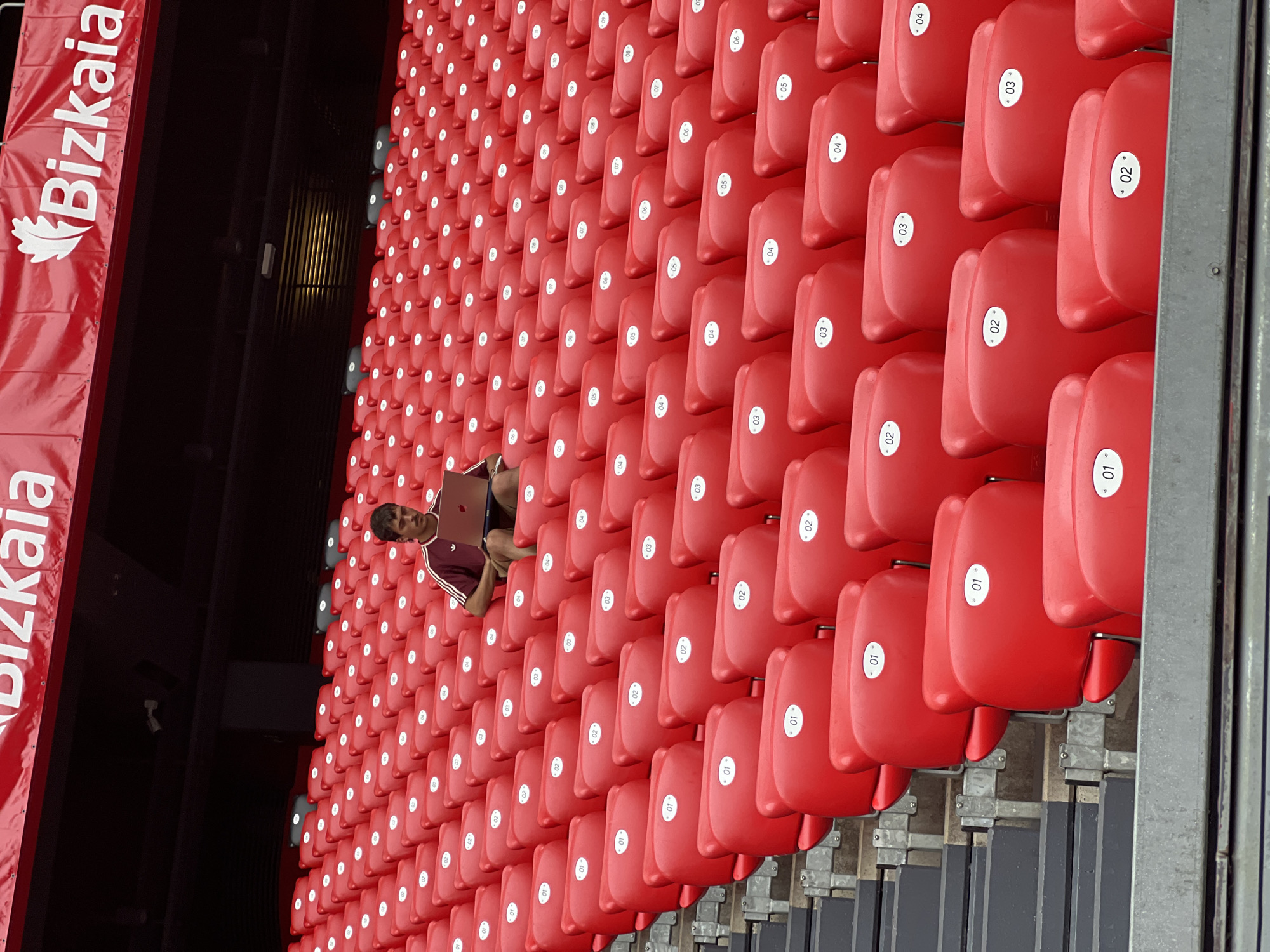 The image shows a young man sitting on red stadium seats, using a silver laptop. He is wearing a burgundy and white striped shirt and shorts. The stadium is filled with rows of identical red seats, many of which are labeled with white numbers. Above the seats, a red banner with white text that reads "Bizkaia" is visible. This suggests the location is in Bilbao, Spain, as "Bizkaia" is the Basque name for the province where Bilbao is located, and the banner likely signifies a sporting or cultural event associated with the region. The stadium appears to be part of San Mamés Stadium, the home ground of Athletic Bilbao football club. The environment is indoors or covered, with dark ceilings and lighting visible in the upper portions of the stadium. The man is positioned in the middle rows of the stadium, with a clear view of the seats extending upwards and outwards.