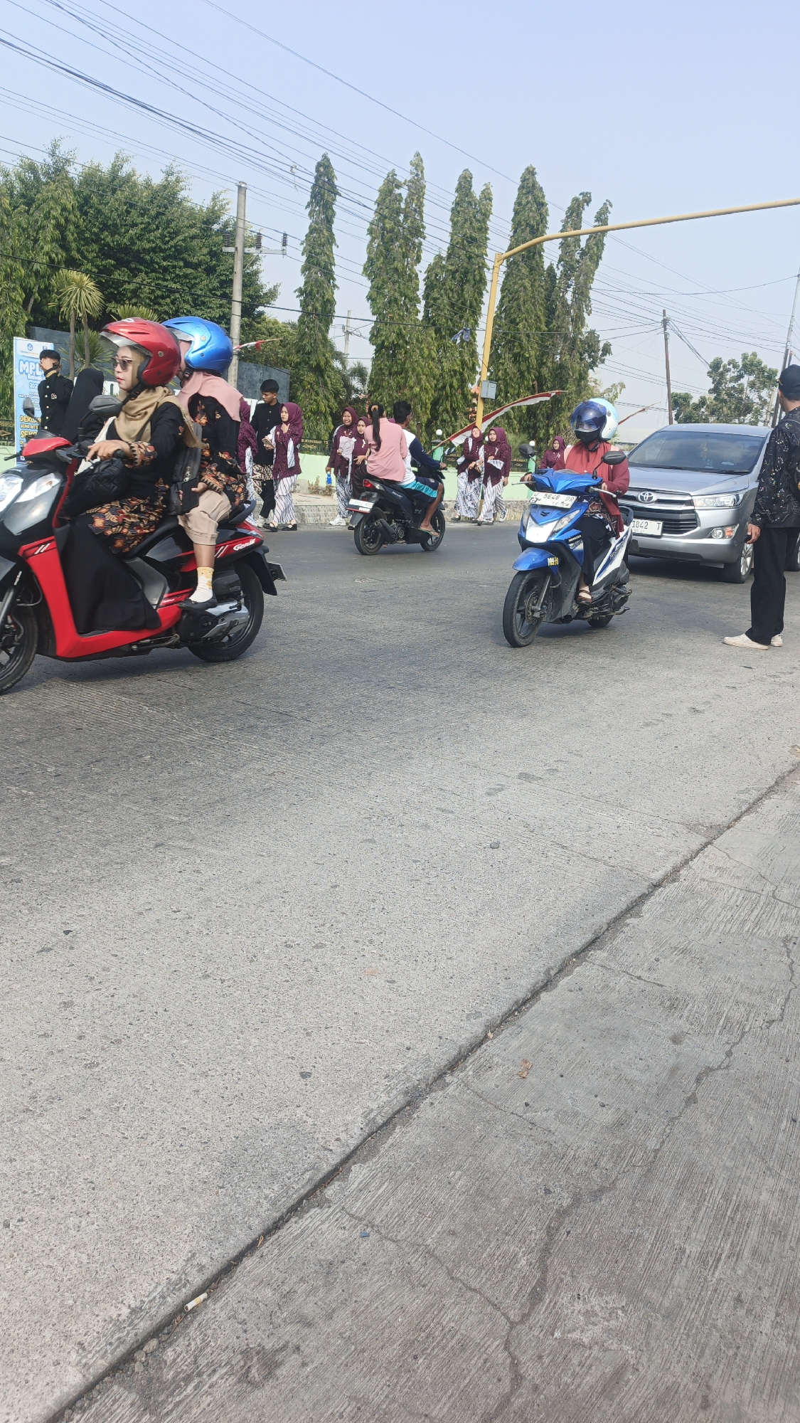 In Sumengko, Indonesia, on a sunny day, a group of people are gathered near a road. Several individuals are riding motorcycles, including a woman in a red helmet and a black and red patterned outfit, a woman in a blue helmet and a pink headscarf, and a person in a red jacket and blue helmet. A silver car is parked on the right side of the road. In the background, a line of tall, slender trees stands against a clear blue sky. A man in a black jacket and dark pants stands on the sidewalk, facing away from the camera. The road is paved with asphalt and shows some cracks and wear.